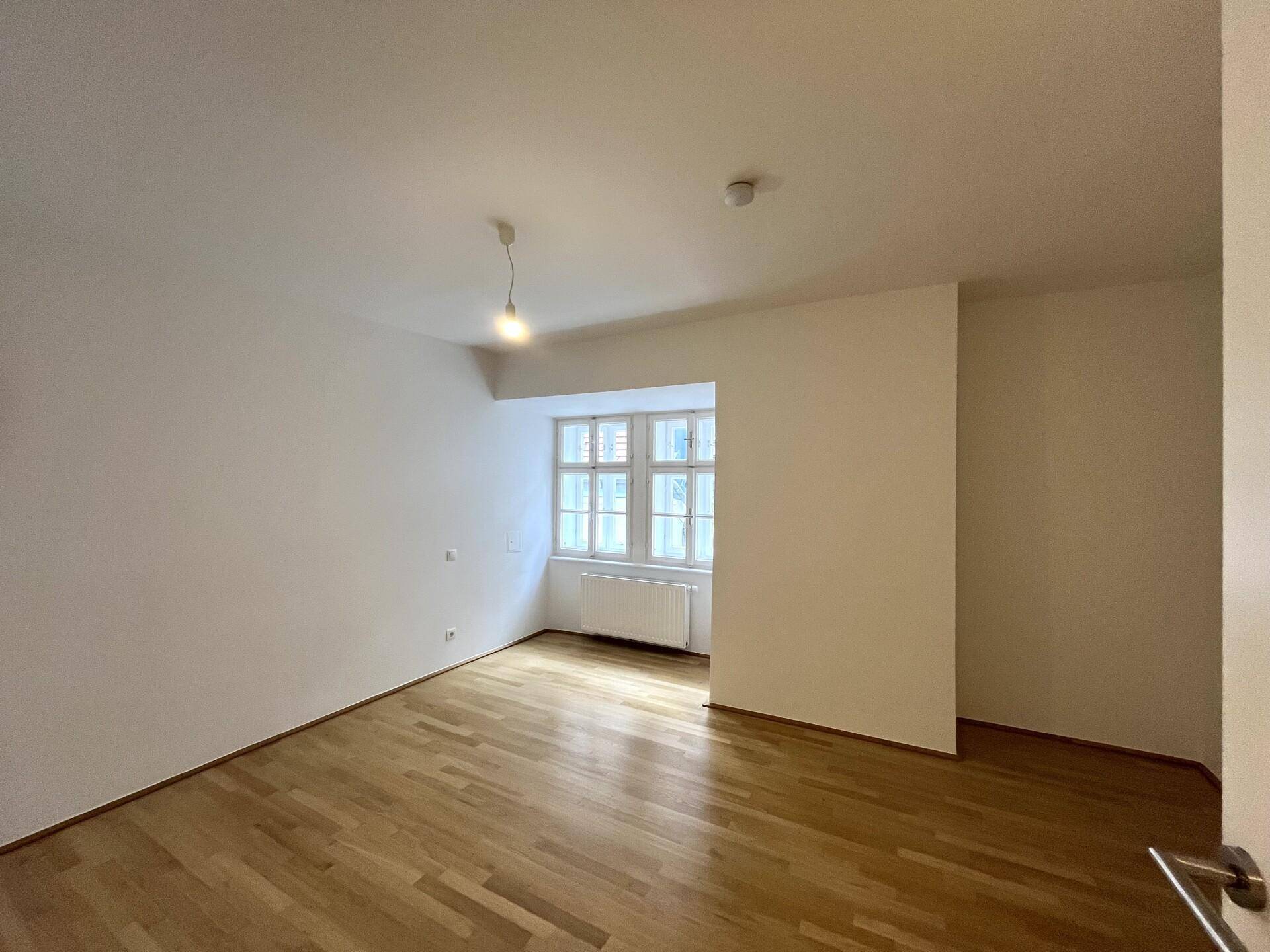 Helles Schlafzimmer mit Parkettboden und einem Fenster, das viel Tageslicht hereinlässt, bereit zur Einrichtung.