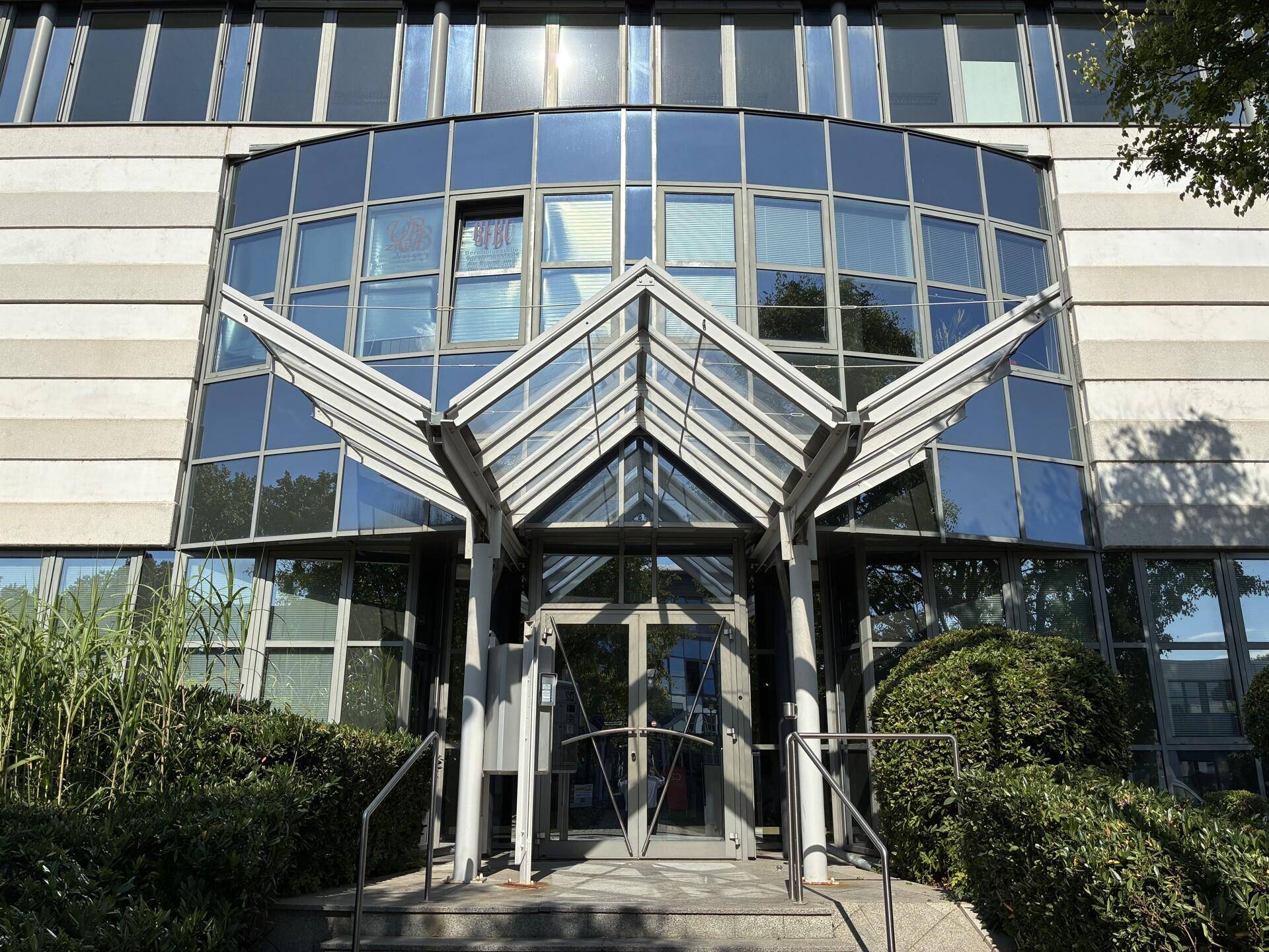 Moderne Bürogebäude-Fassade mit Glasvordach und großen Fenstern, umgeben von Grünflächen.