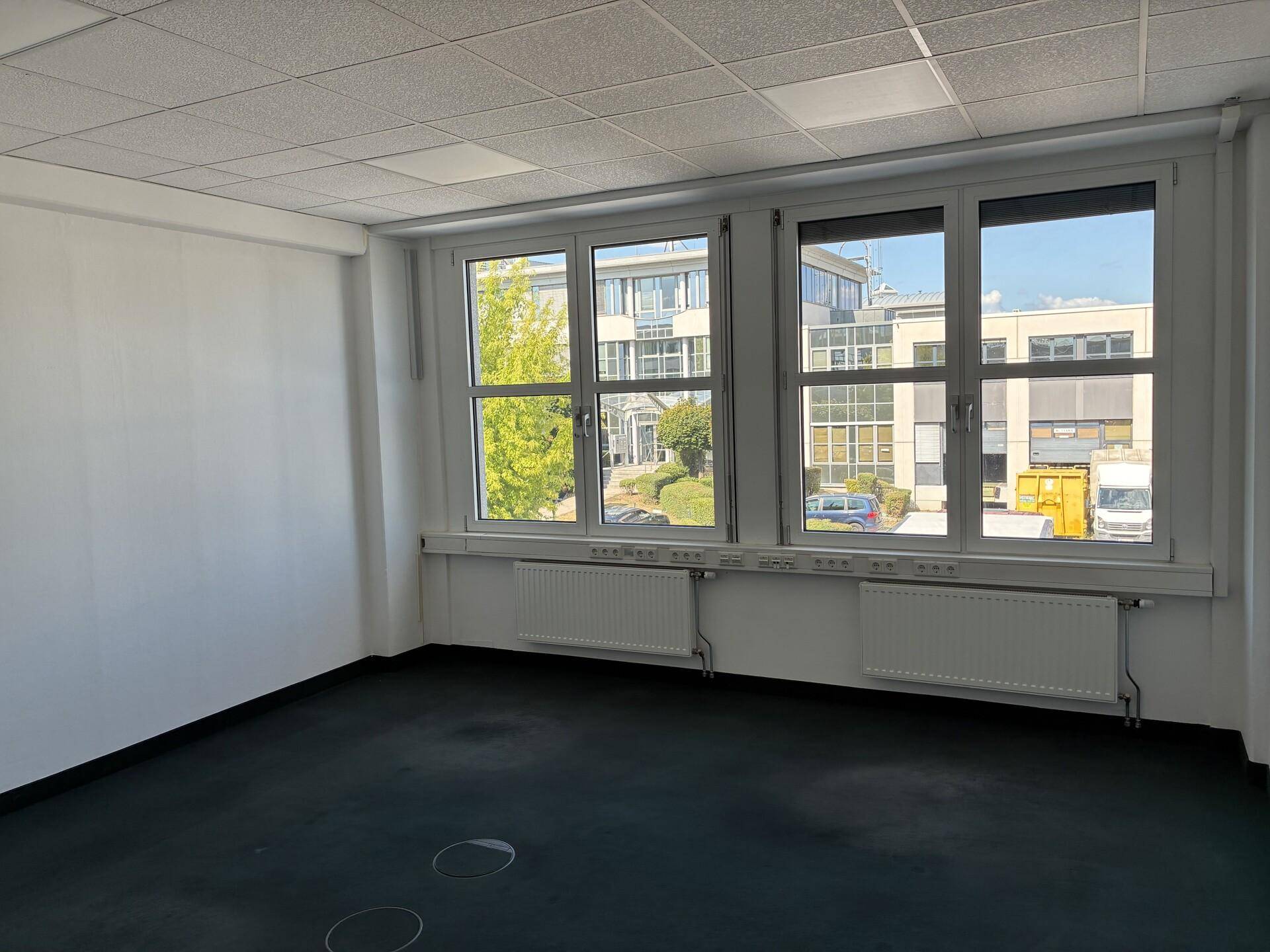 Helles Büro mit Fenstern, die auf ein gegenüberliegendes Gebäude und einen Parkplatz blicken, mit dunklem Teppich.