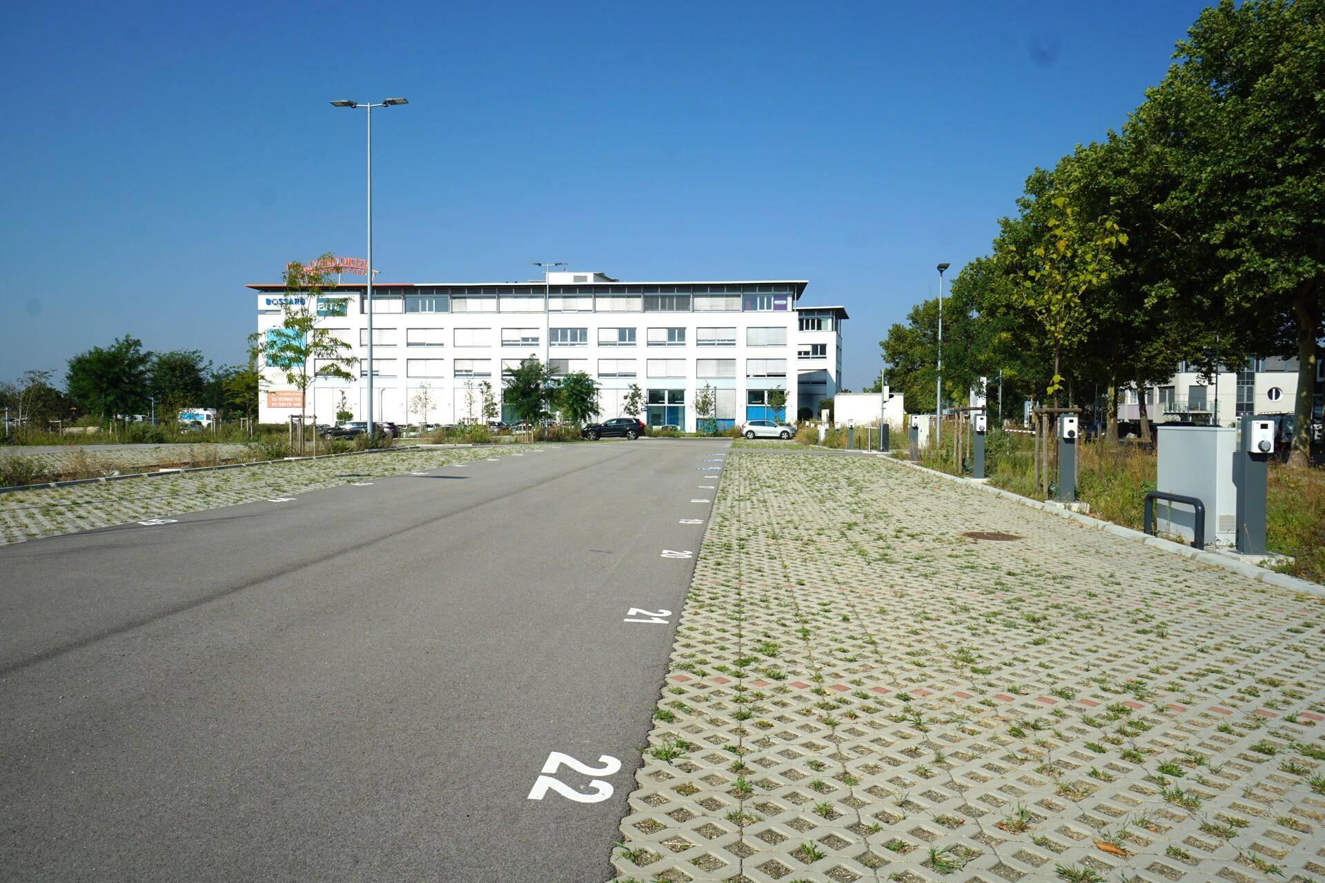 Großer Parkplatz mit mehreren Ladestationen für Elektrofahrzeuge und einem Bürogebäude im Hintergrund.