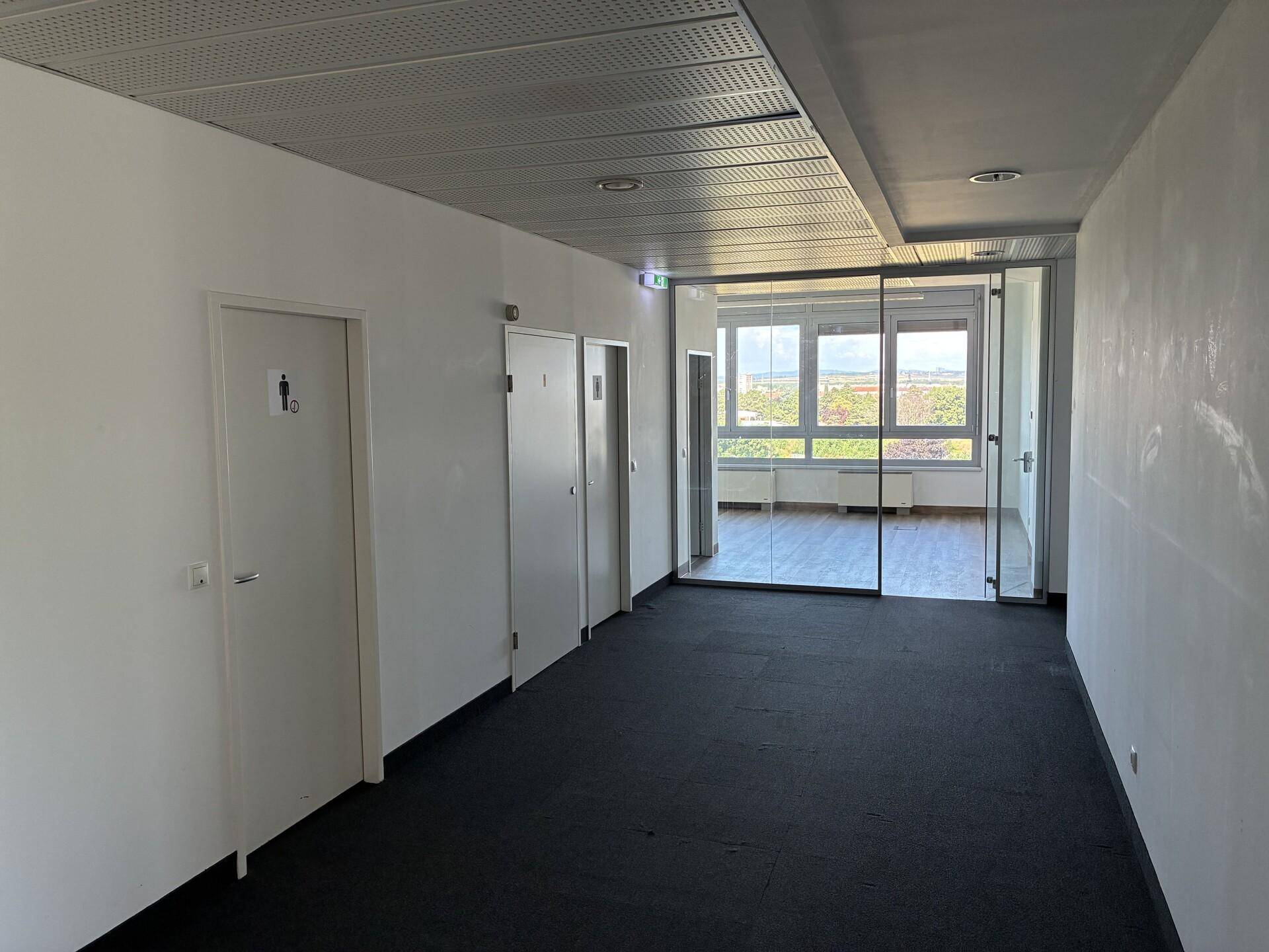 Langer Flur mit Teppichboden, abgehängter Decke und Blick in ein helles Büro.
