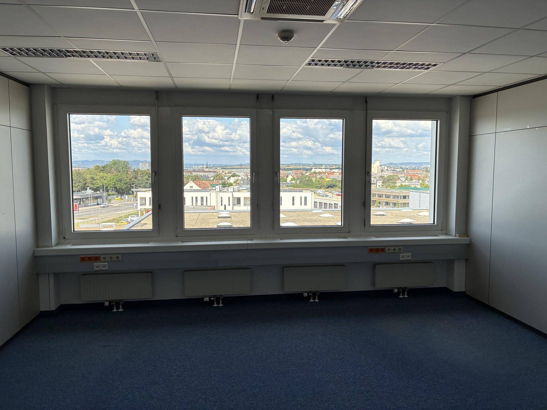 Büro mit breiter Fensterfront, blauem Teppichboden und Blick über die Stadtlandschaft.