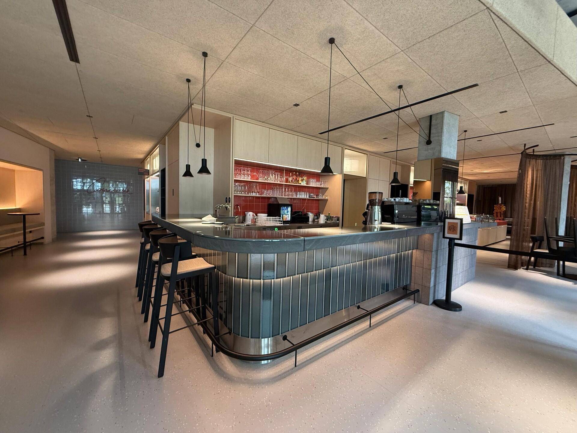 Moderne Bar mit langer Theke, Barhockern und stilvollen Pendelleuchten, ergänzt durch eine rote Fliesenrückwand.