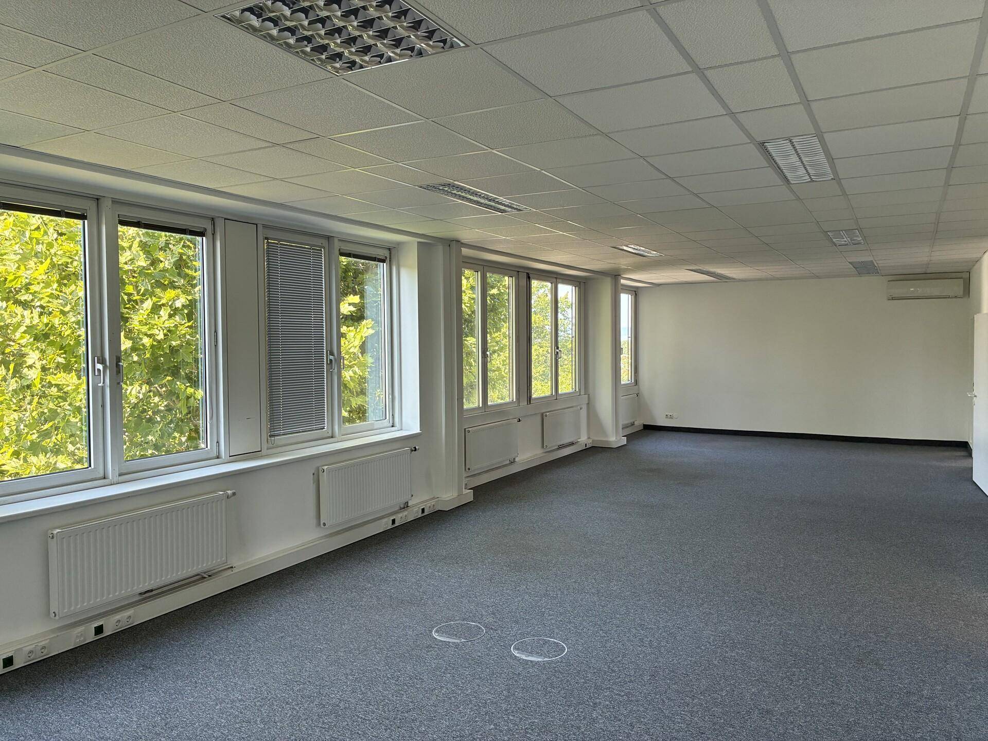 Großer, leerer Bürobereich mit Teppichboden, vielen Fenstern mit Jalousien und Klimaanlage im Hintergrund.