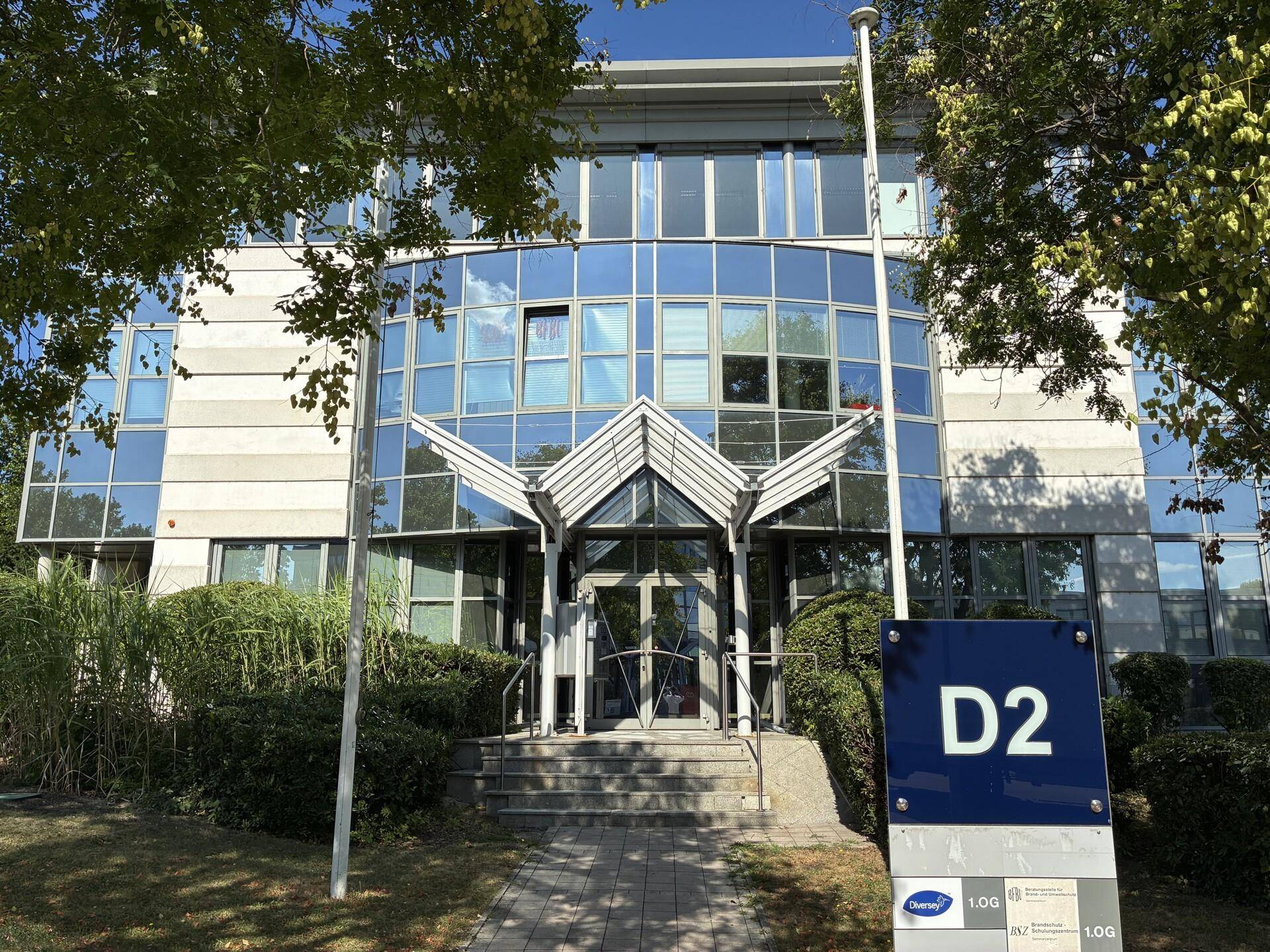Moderne Bürogebäude-Fassade mit viel Glas, umgeben von Bäumen und Sträuchern, mit einem Hinweisschild im Vordergrund.