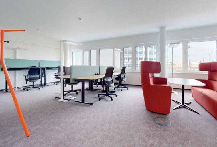 Geräumiges Großraumbüro mit Teppichboden, ergonomischen Arbeitsplätzen und farbigen Lounge-Sesseln.
