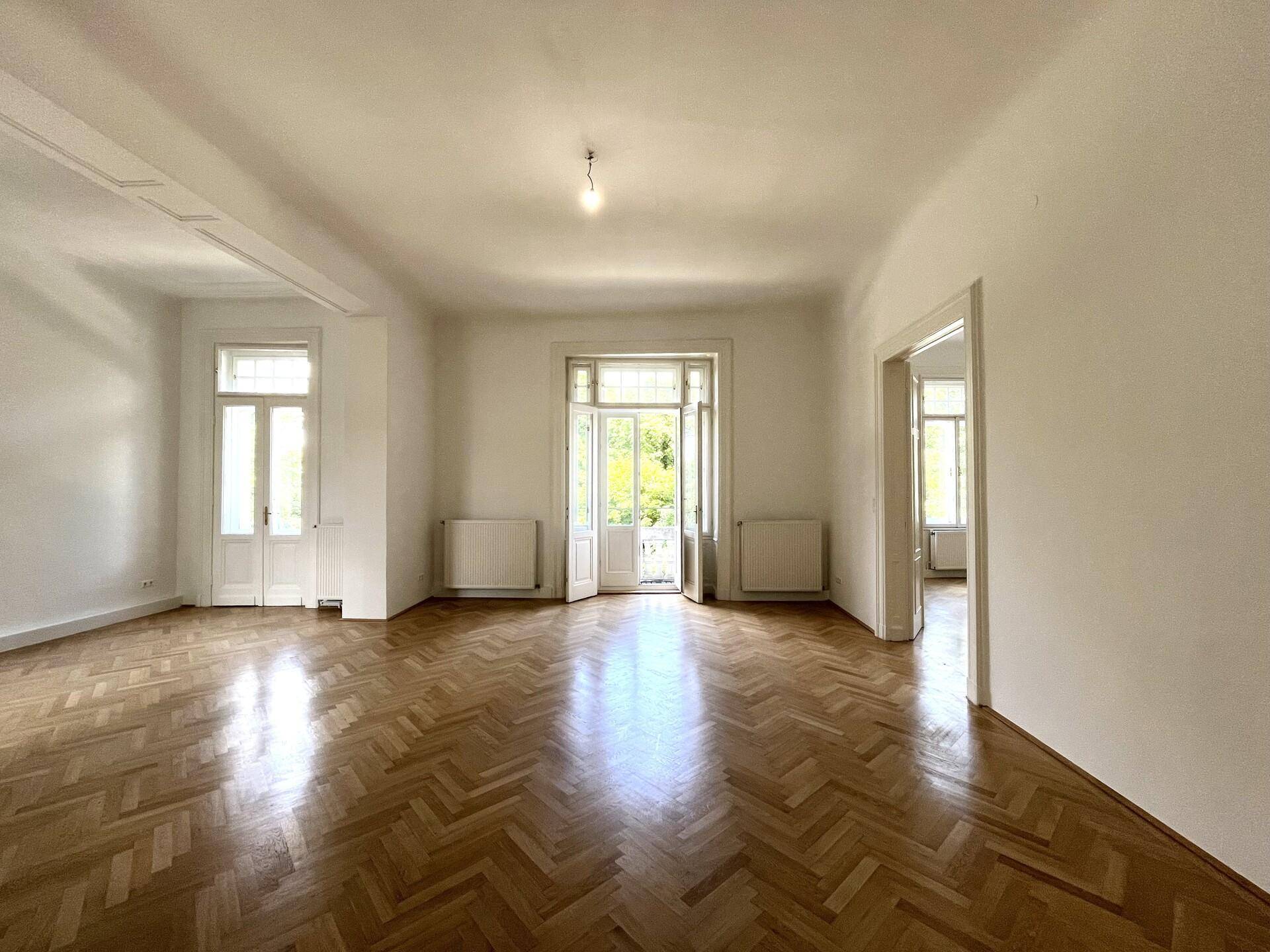 Helles, leeres Zimmer mit Fischgrätparkett und Zugang zu einem Außenbereich durch Flügeltüren.