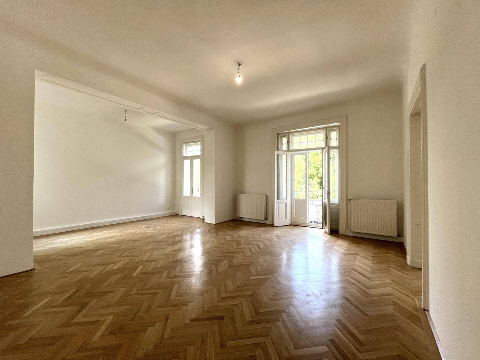 Großes, helles Zimmer mit Fischgrätparkett und hohen Türen, die zu einem Außenbereich führen.