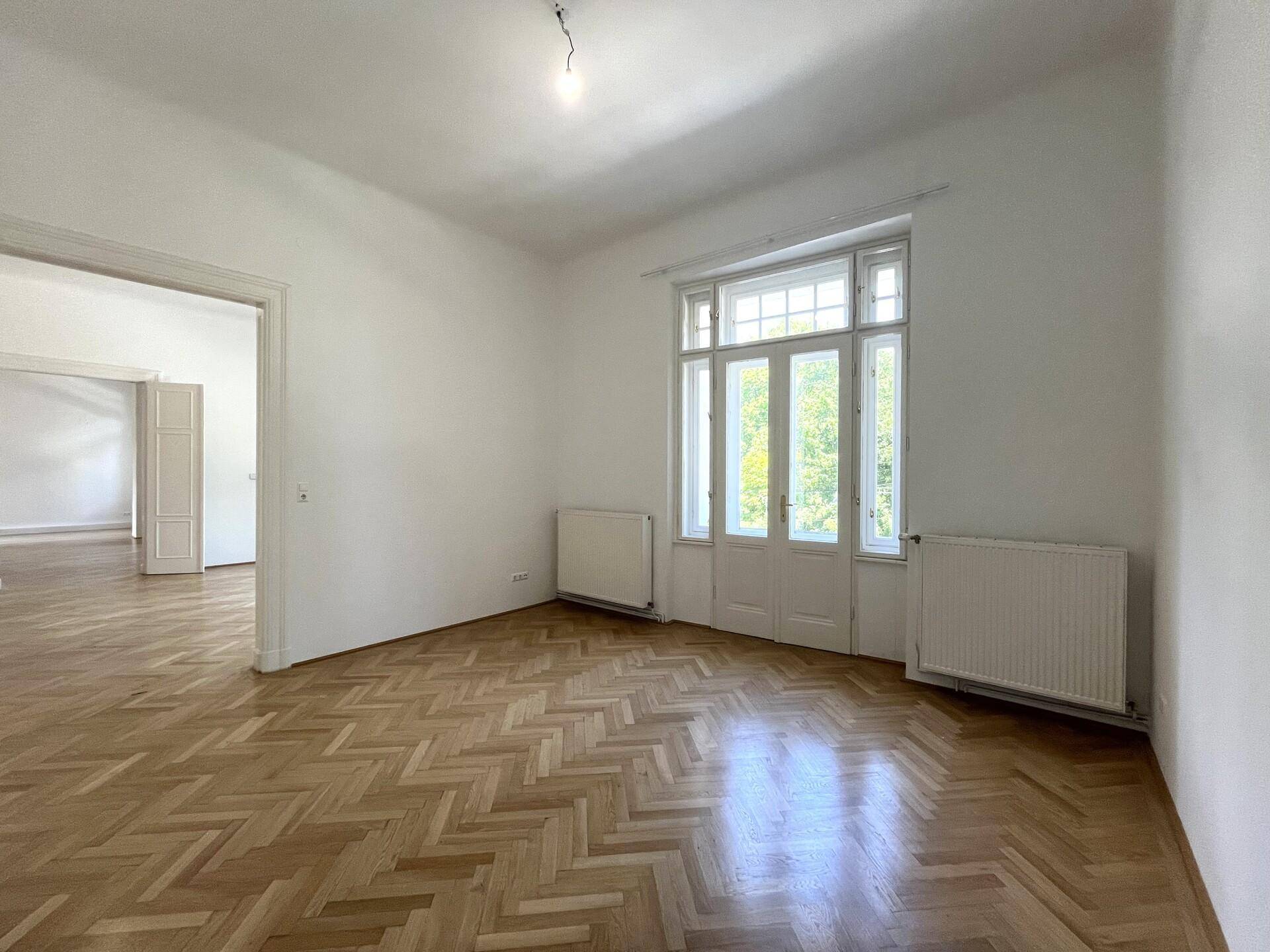 Leeres Zimmer mit glänzendem Fischgrätparkett und einem hohen Fenster, das viel Licht hereinlässt.