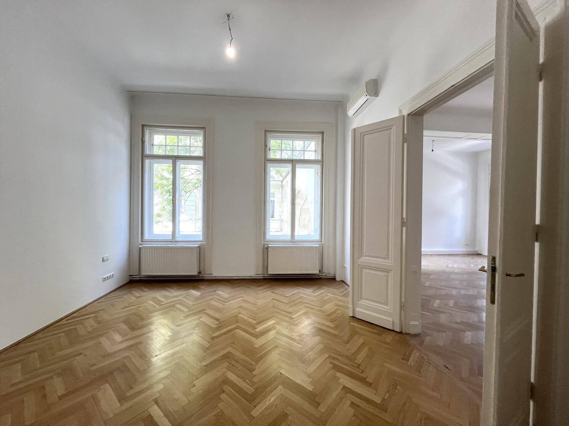 Helles Zimmer mit Fischgrätparkett, zwei Fenstern und einer Klimaanlage an der Wand.