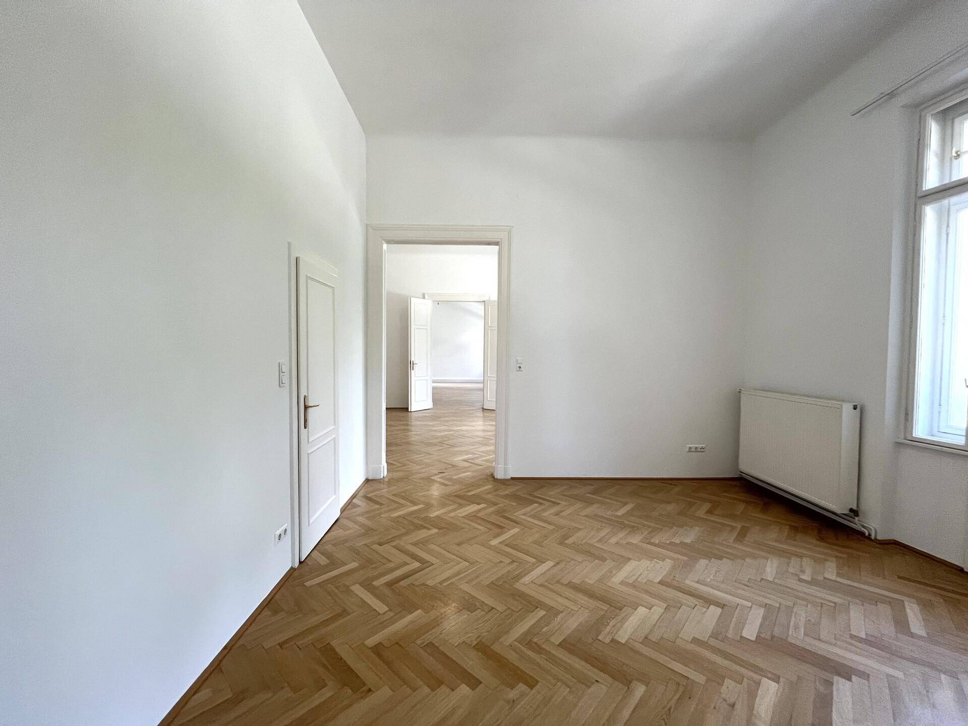 Helles, leeres Zimmer mit Fischgrätparkett und offenem Durchgang zu einem weiteren Raum.