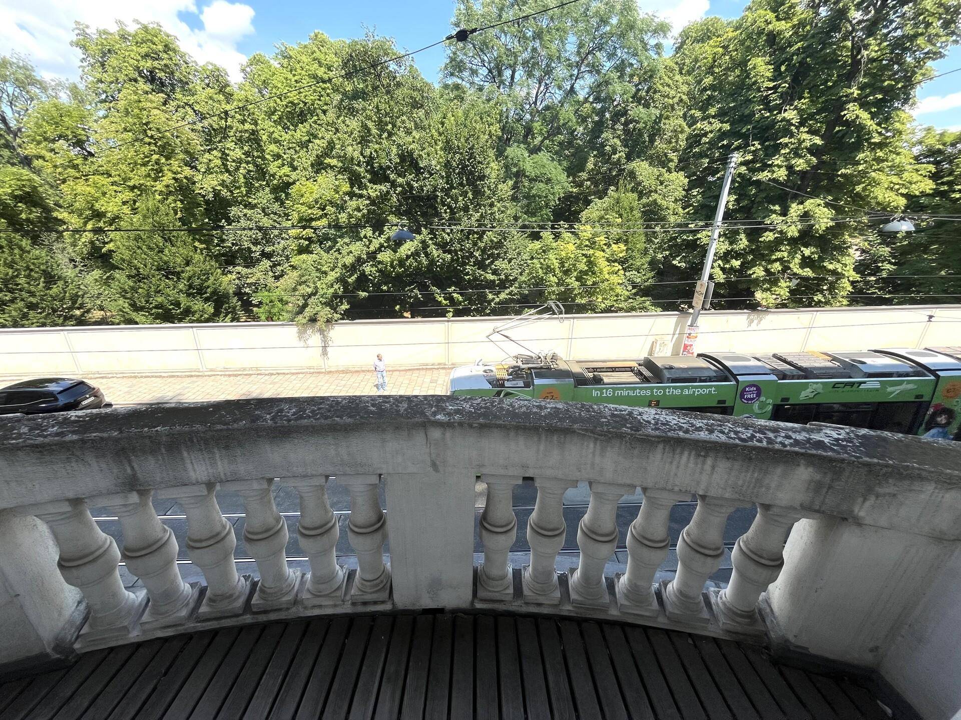Ausblick von einem Balkon mit Balustrade auf eine belebte Straße mit Straßenbahn und Bäumen.