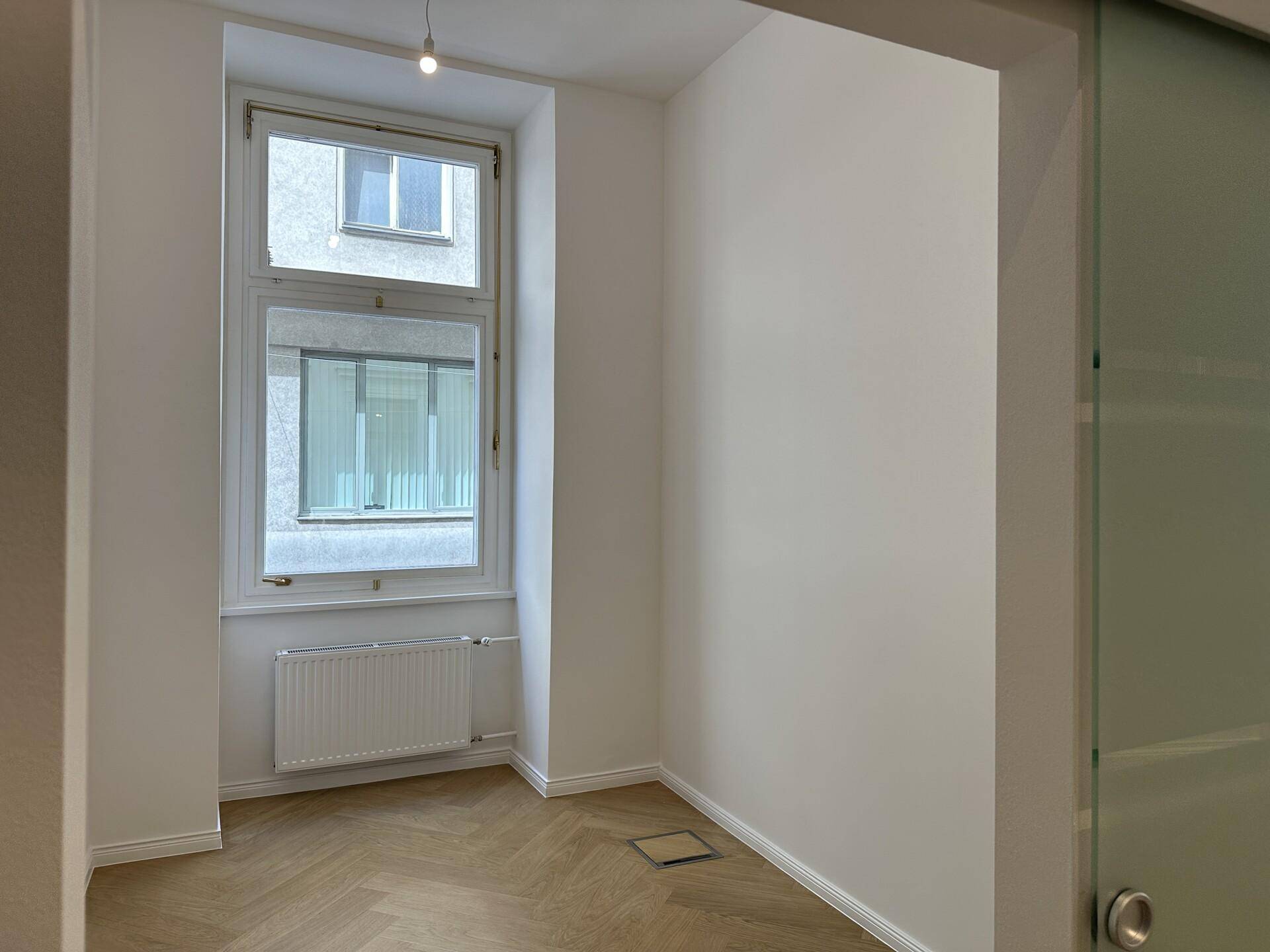 Helles Zimmer mit Fischgrätparkett, einem großen Fenster und einer modernen Schiebetür.
