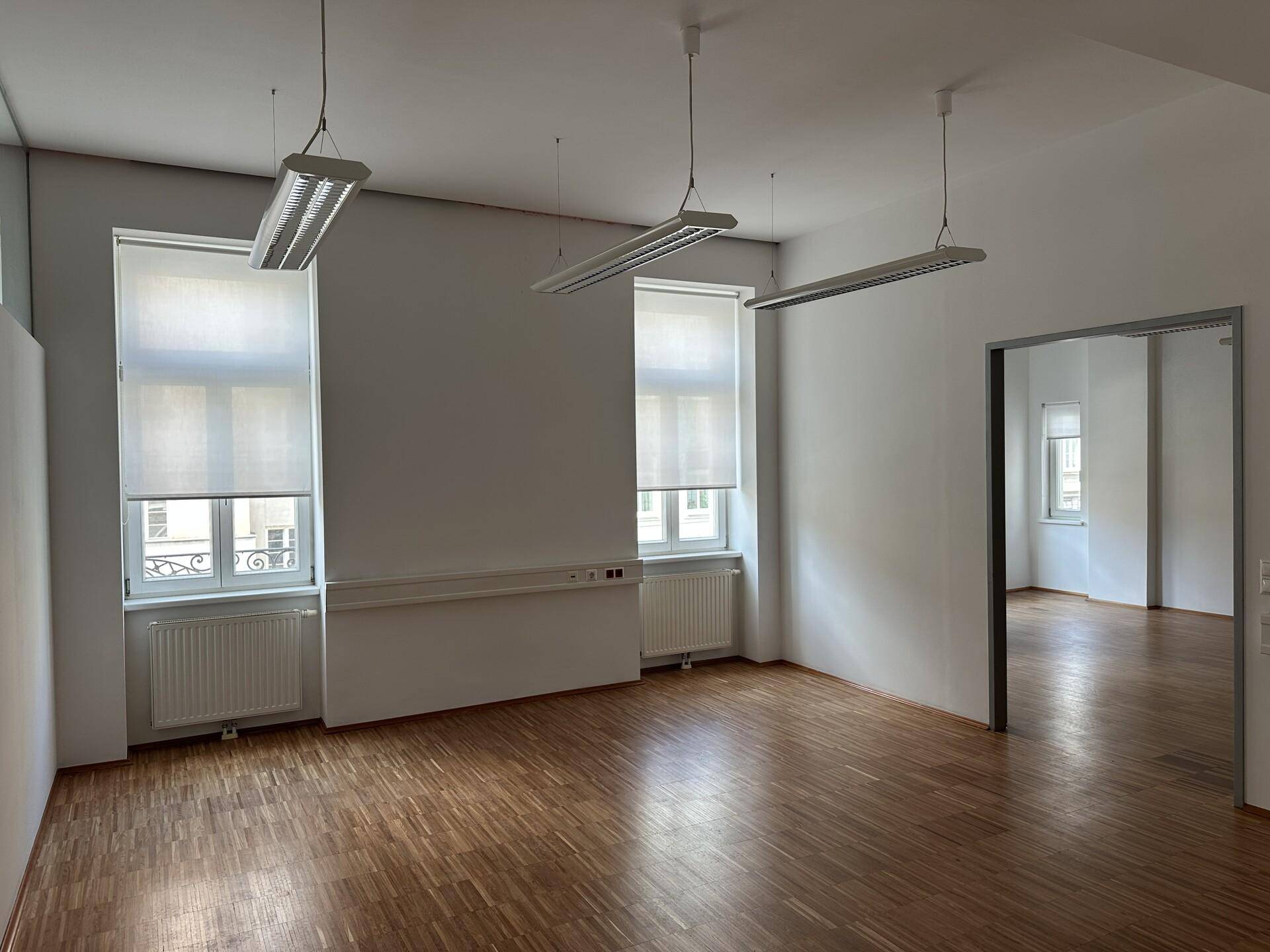 Helles Büro mit Holzboden, zwei Fenstern und einer offenen Tür zu einem weiteren Raum.