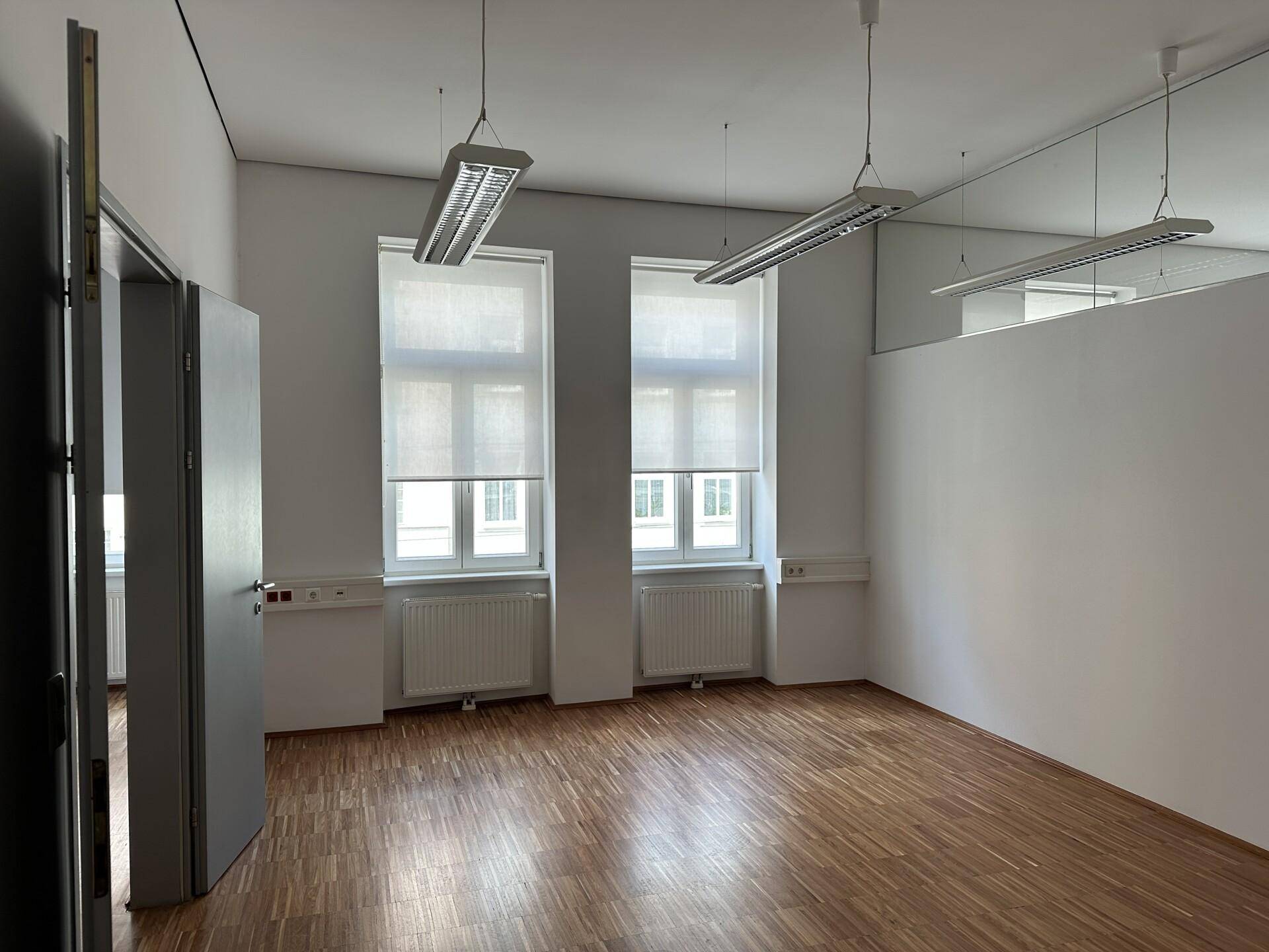 Helles Büro mit Holzparkettboden, großen Fenstern mit Rollos und funktionaler Deckenbeleuchtung.