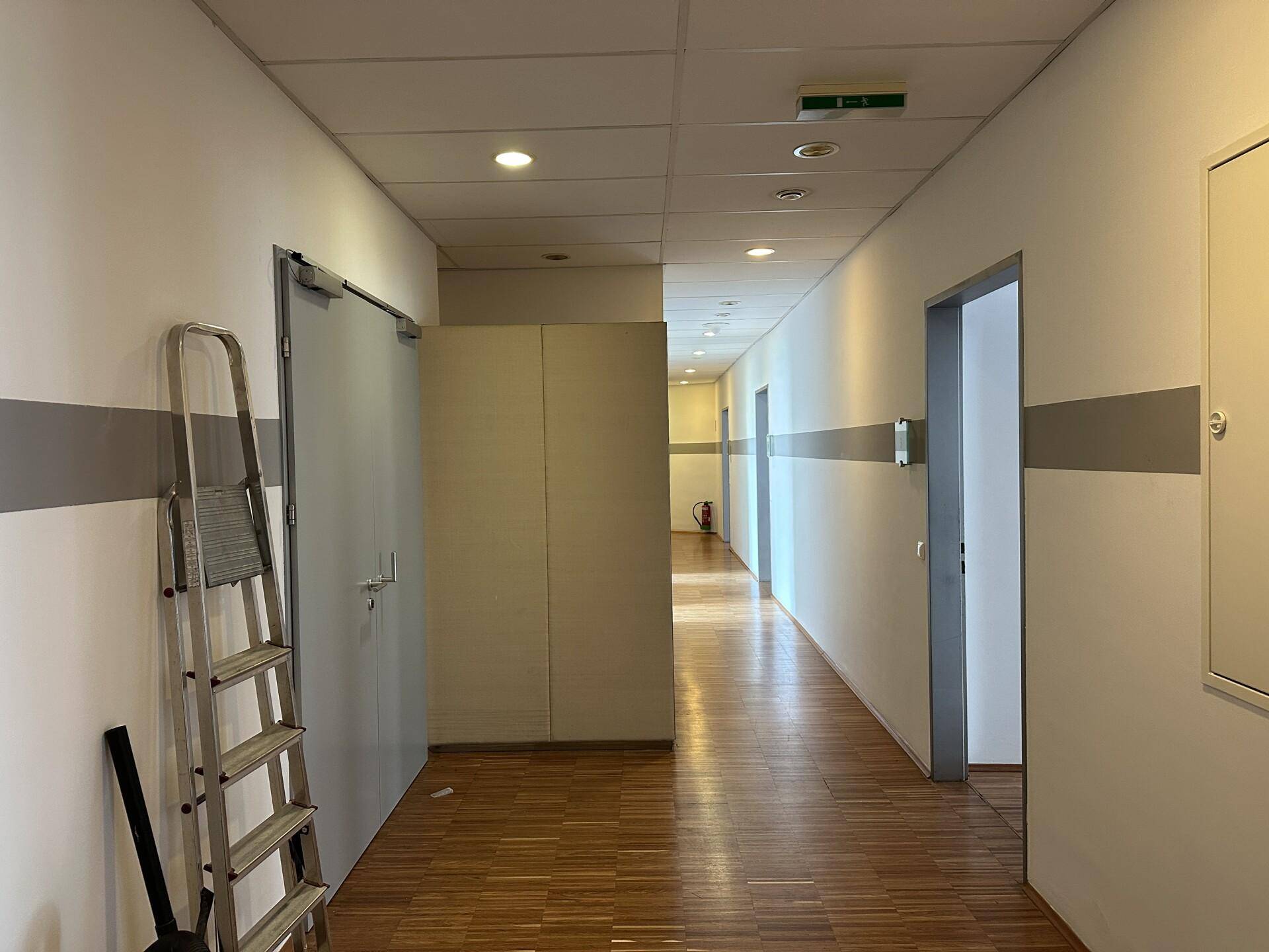 Langer Flur mit Holzboden, weißen Wänden und Deckenleuchten, führt zu mehreren Türen.