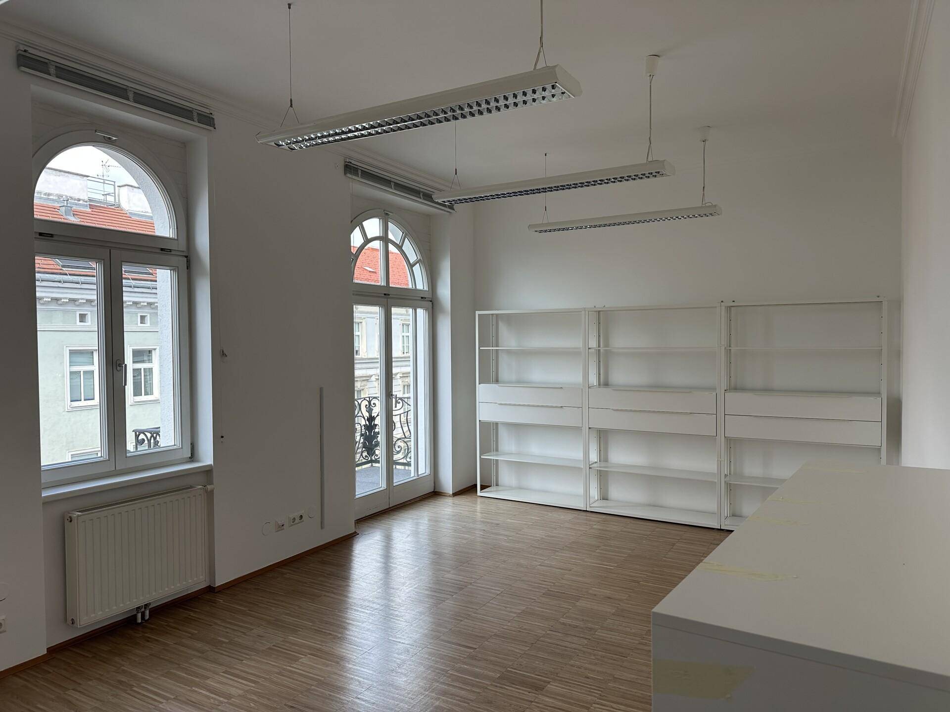 Geräumiges Büro mit hohen Bogenfenstern, Holzboden und weißen Regalen an der Wand.