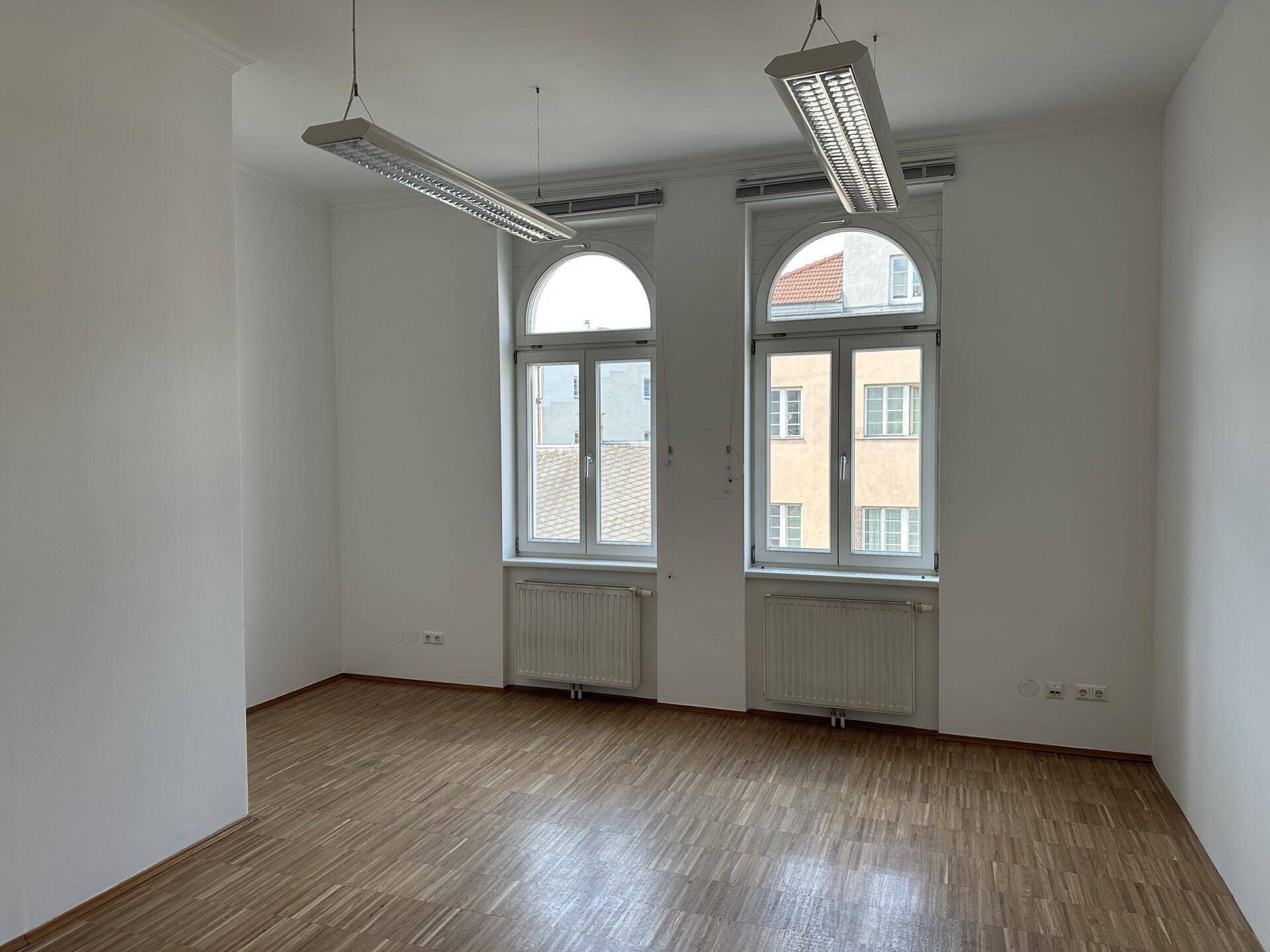 Helles Büro mit zwei hohen Bogenfenstern, Holzboden und Deckenleuchten.