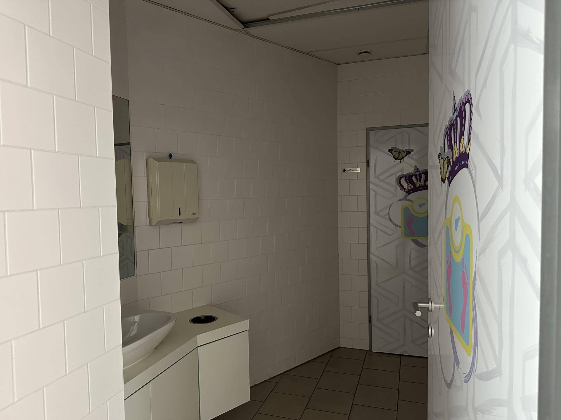 Badezimmer mit weißen Fliesen, modernem Waschbecken und einer Tür mit farbenfroher Gestaltung.