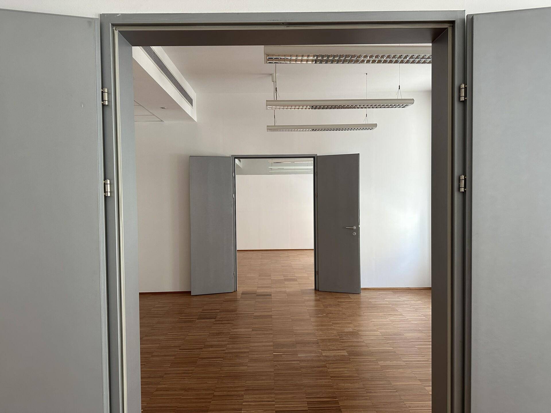 Blick durch eine offene Tür in einen weiteren leeren Raum mit Holzboden und hohen Decken.