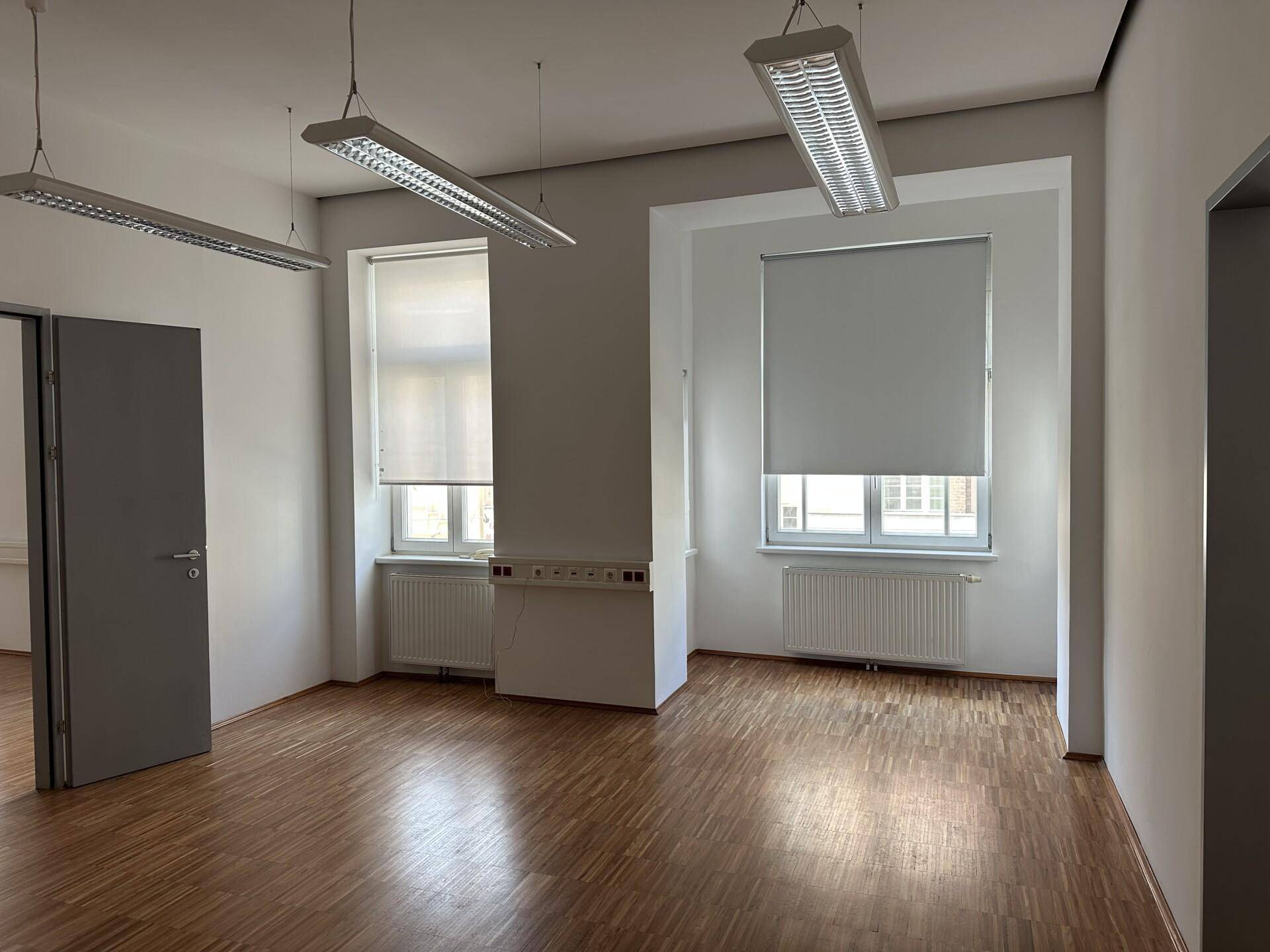 Geräumiger Büroraum mit Holzboden, weißen Wänden und zwei Fenstern, bereit zur Einrichtung.