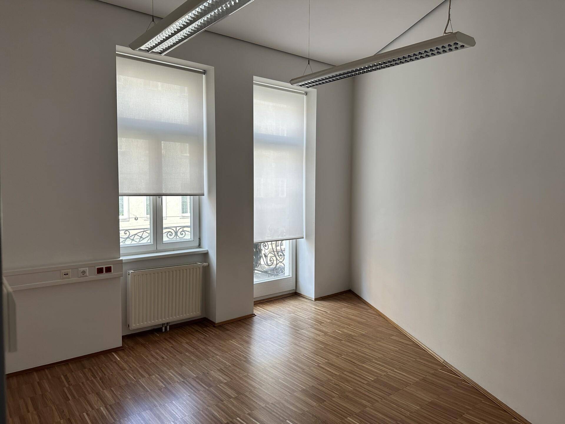 Heller Büroraum mit Holzboden, weißen Wänden und zwei Fenstern, eines davon mit französischem Balkon.