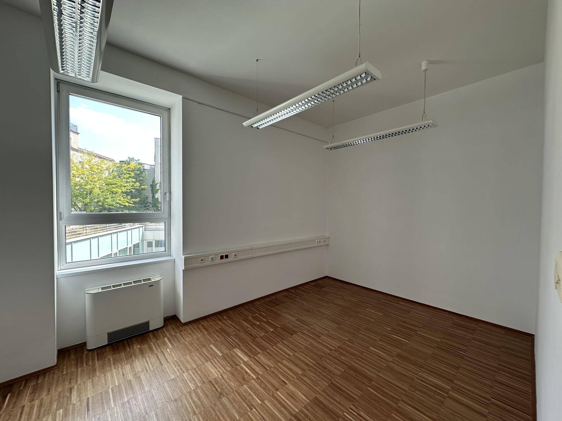 Leerer Büroraum mit großem Fenster, Holzboden und einer Klimaanlage, die viel Tageslicht bietet.