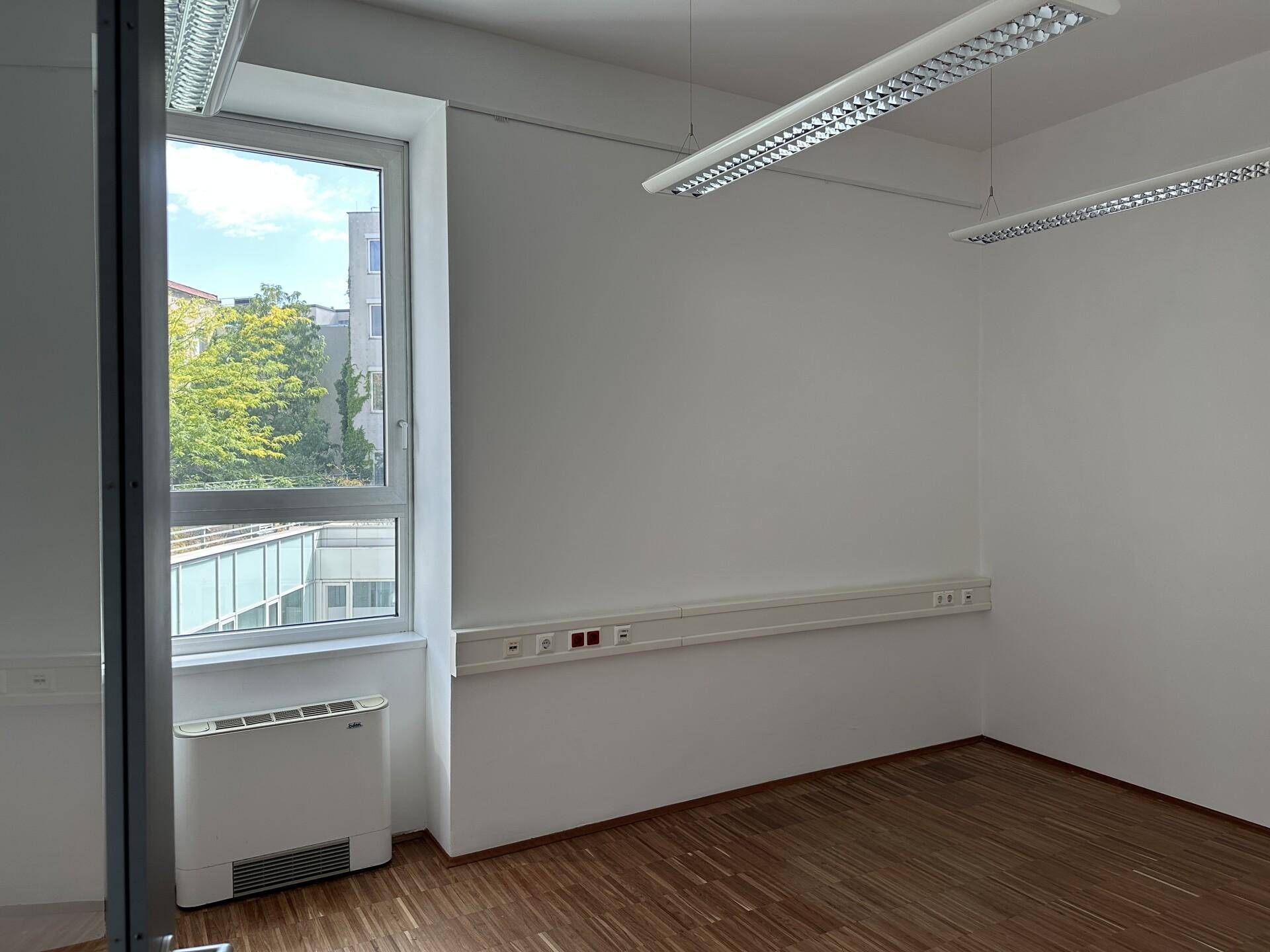 Heller Büroraum mit großem Fenster, Holzboden und Klimaanlage, ideal für produktives Arbeiten.