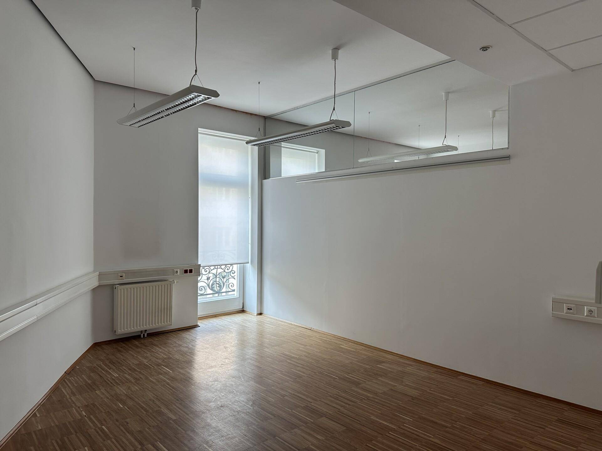 Geräumiger Büroraum mit Holzboden, weißen Wänden und einem Fenster mit Blick nach draußen.