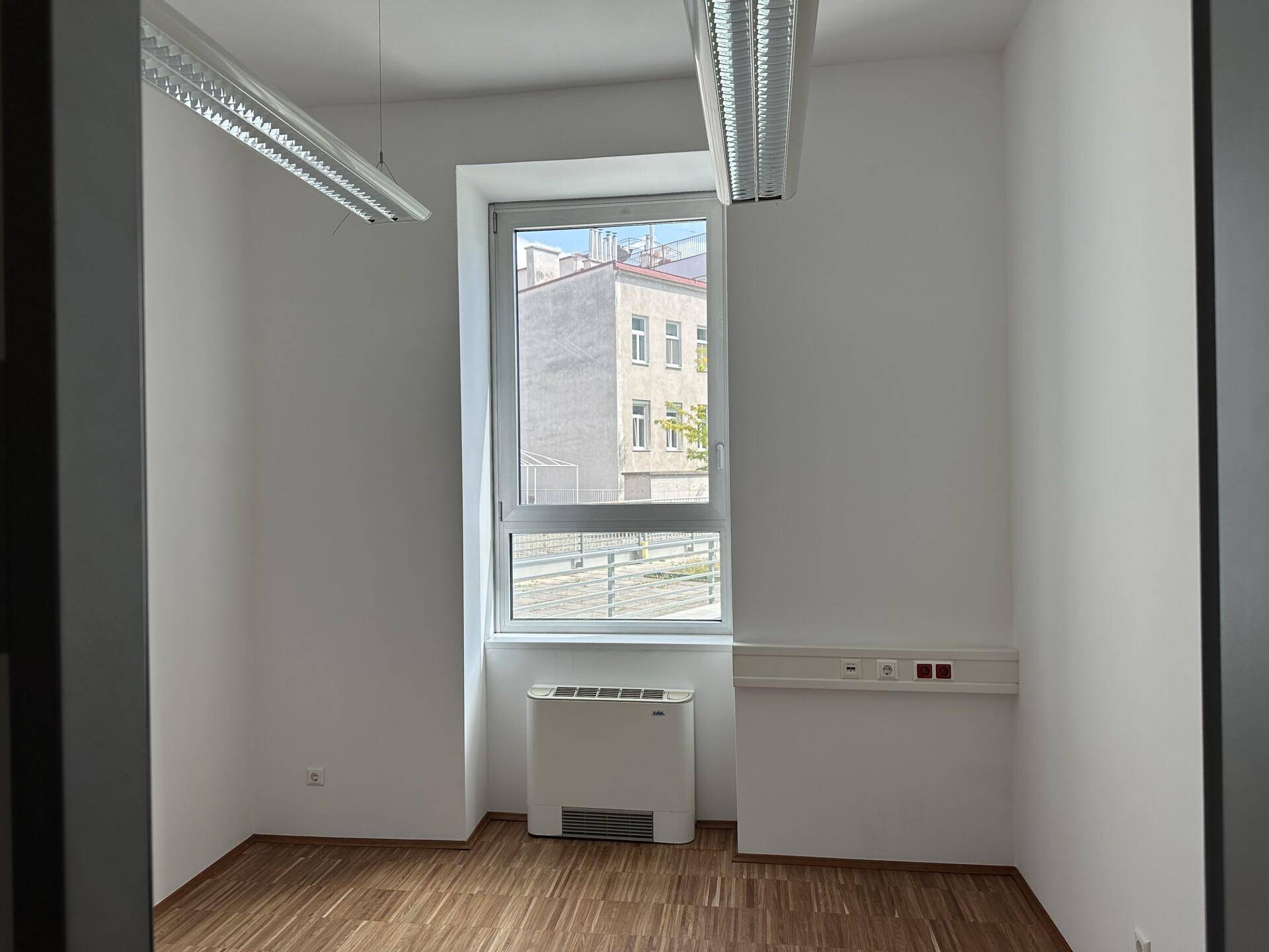 Leerer Büroraum mit Holzboden, weiß gestrichenen Wänden und einem großen Fenster mit Stadtblick.