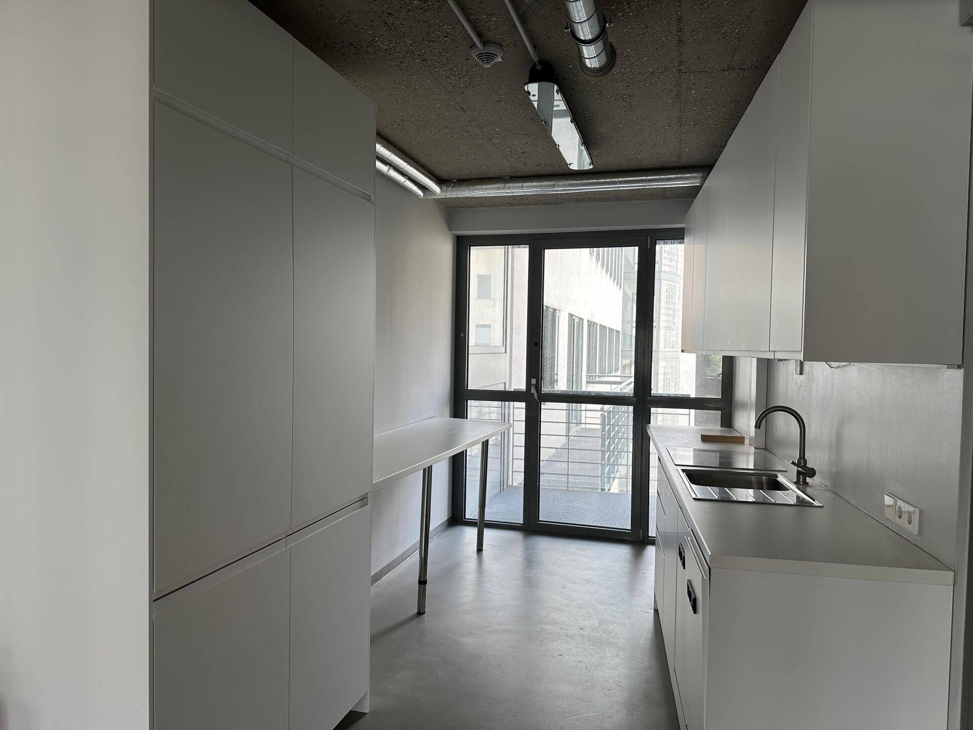 Schmale Küchenzeile mit weißen Schränken, Spüle und Blick auf die Stadt durch große Fenster.