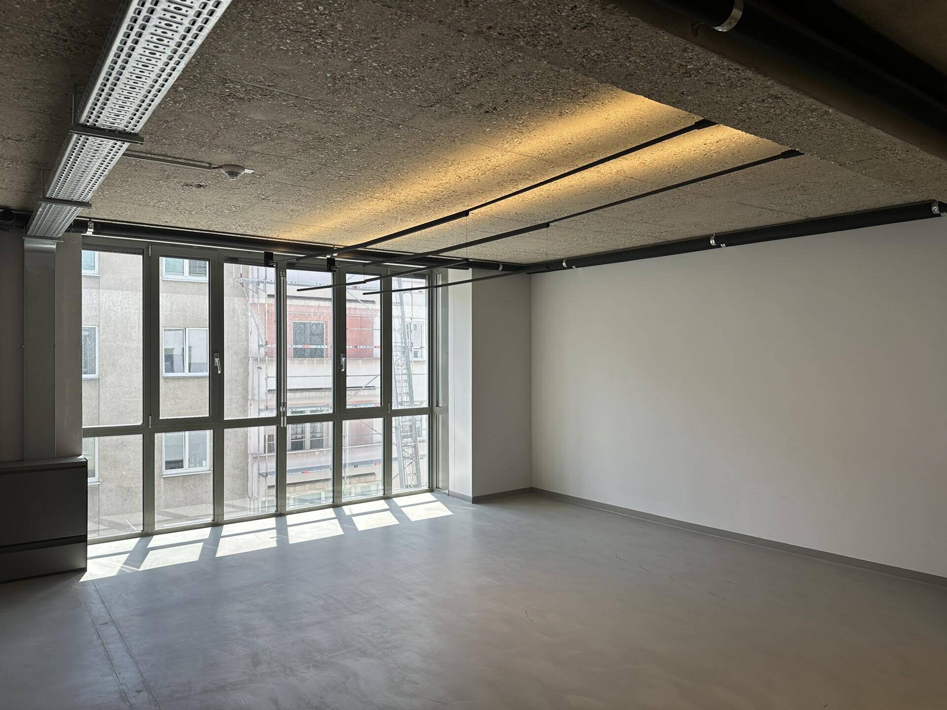 Eckbüro mit großen Fenstern, die viel Sonnenlicht hereinlassen, und einer modernen Sichtbetondecke.