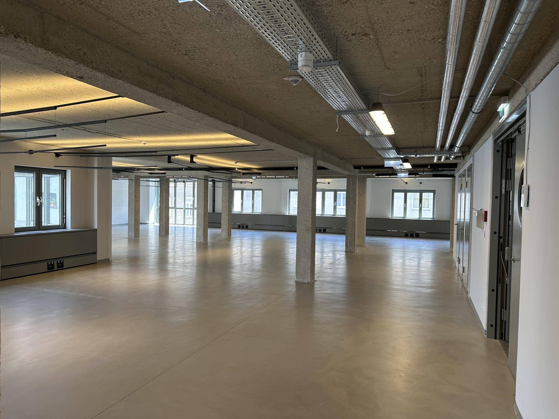 Geräumiger, offener Bürobereich mit vielen Fenstern, Betonsäulen und industrieller Deckeninstallation.