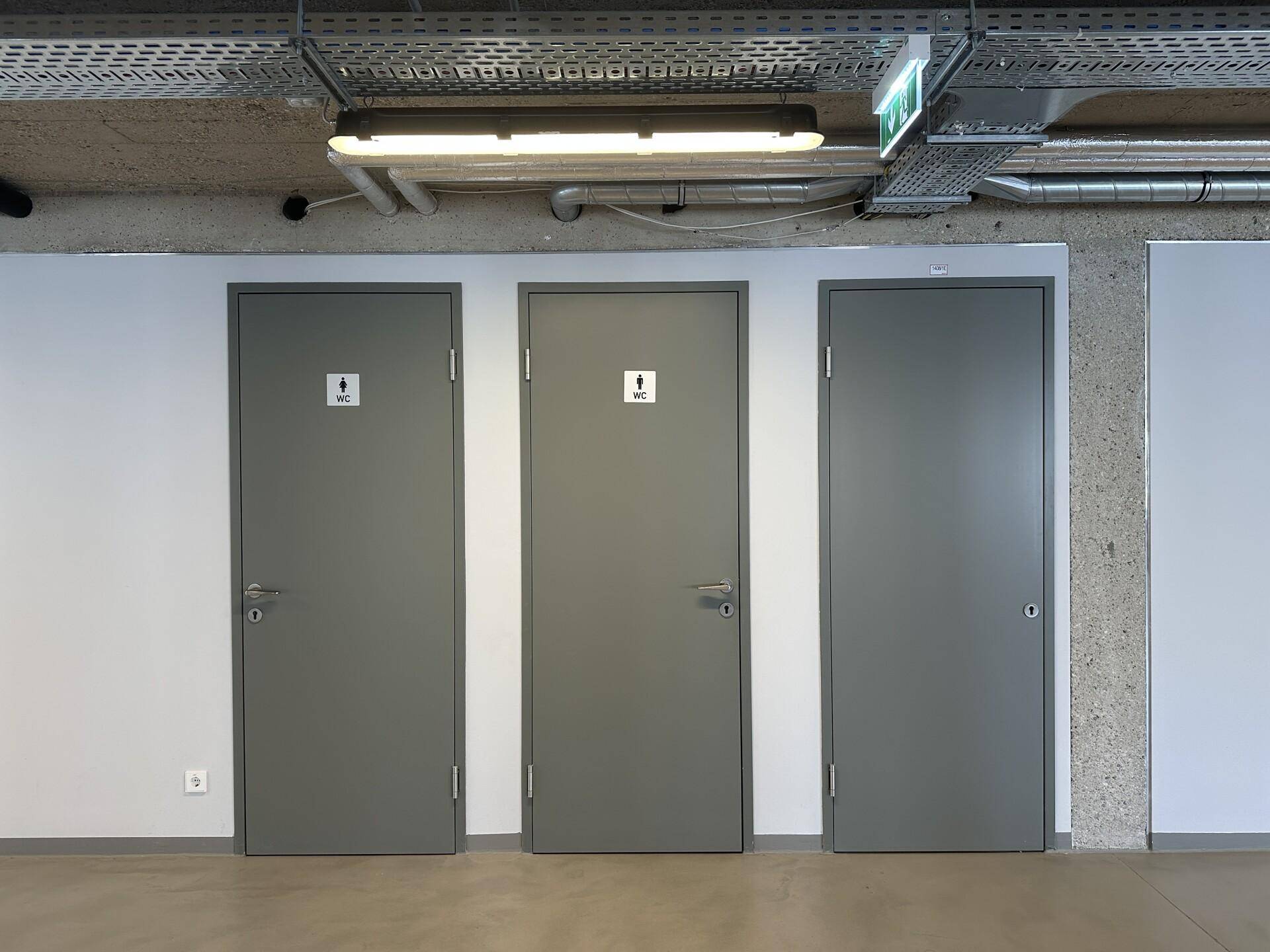 Drei moderne Toilettentüren in einem Flur mit industrieller Deckenbeleuchtung und Beton.