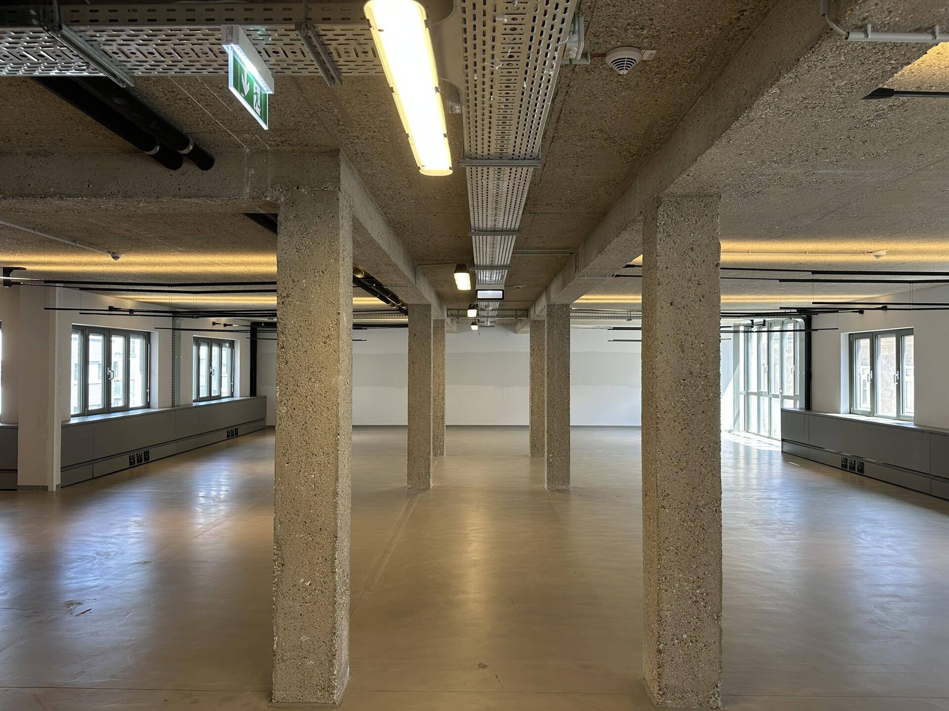 Weitläufiger, offener Bürobereich mit Betonsäulen, industrieller Decke und vielen Fenstern.