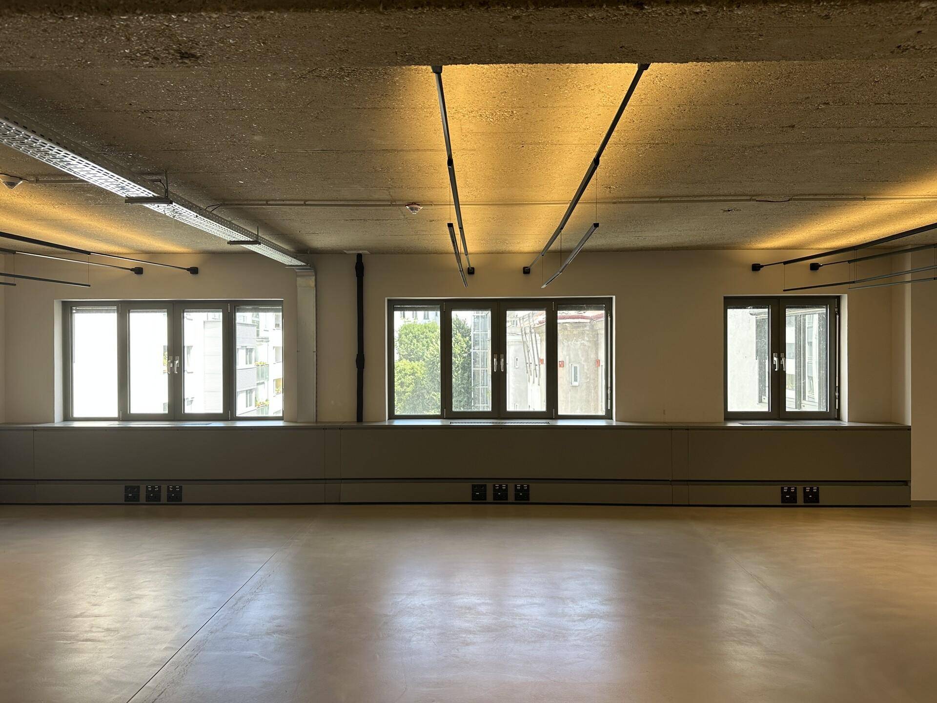 Heller Büroraum mit großen Fenstern, die viel Tageslicht hereinlassen, und einer Sichtbetondecke.