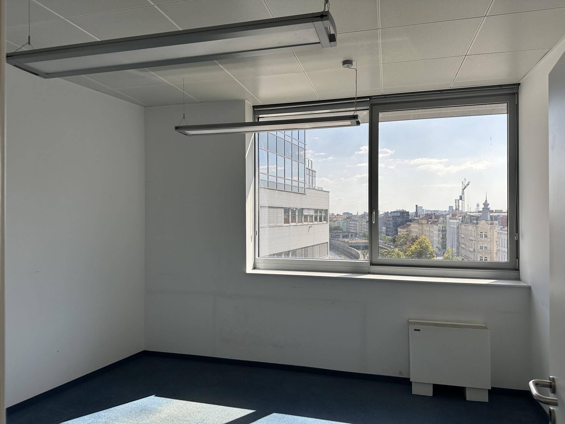 Kleines Büro mit weitem Stadtblick auf moderne Gebäude und blauem Teppichboden.