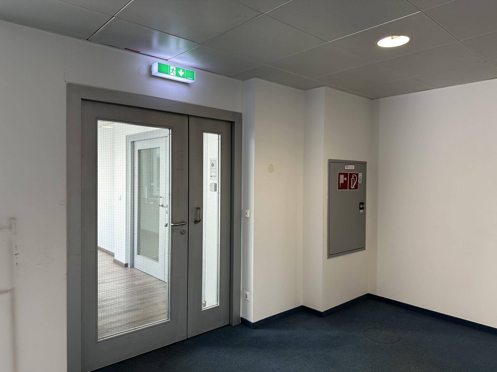 Eingangsbereich mit Doppeltüren, Notausgangsbeschilderung und einem Feuerlöscherschrank an der Wand.