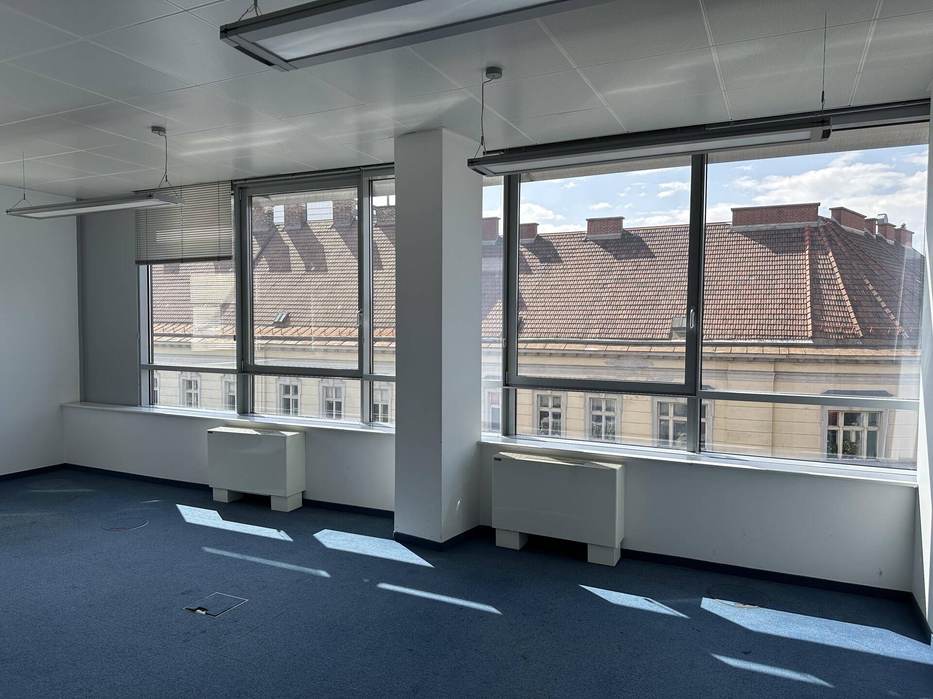 Helles Büro mit blauer Auslegware, großen Fenstern und Blick auf die umliegenden Gebäude.
