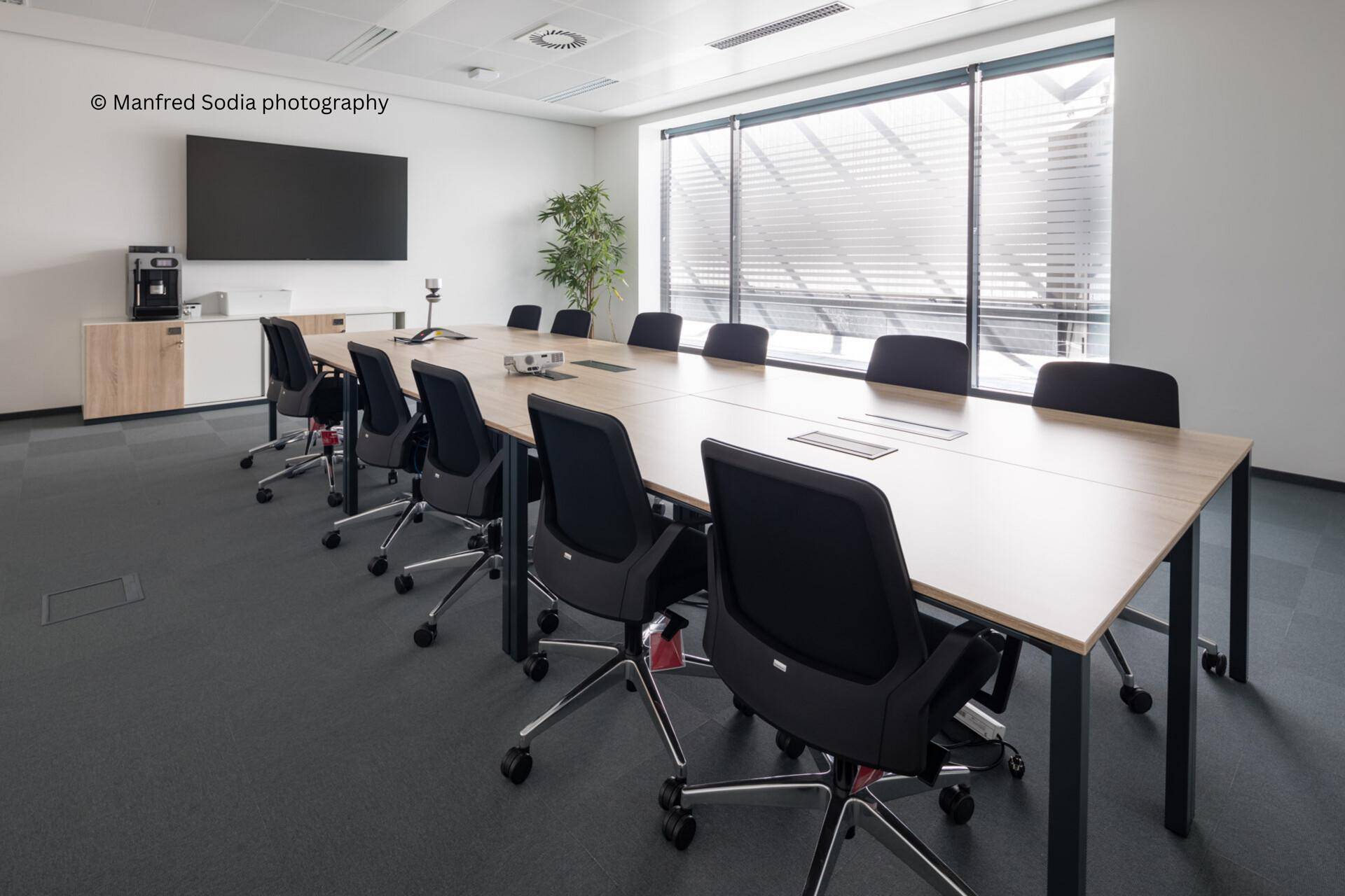 Heller Konferenzraum mit großem Holztisch, modernen Stühlen und einem Flachbildschirm an der Wand.
