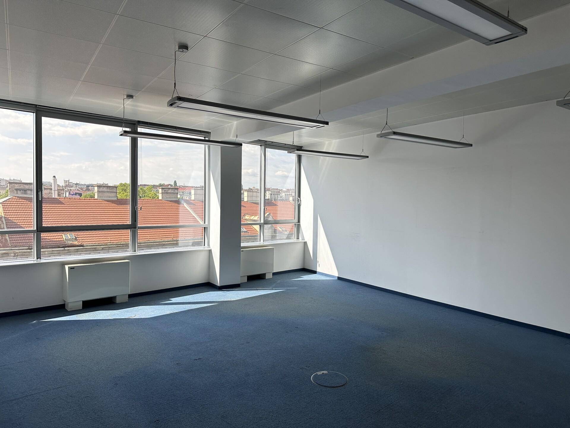 Geräumiges, leeres Büro mit großen Fenstern, blauem Teppichboden und Blick auf die Dächer.