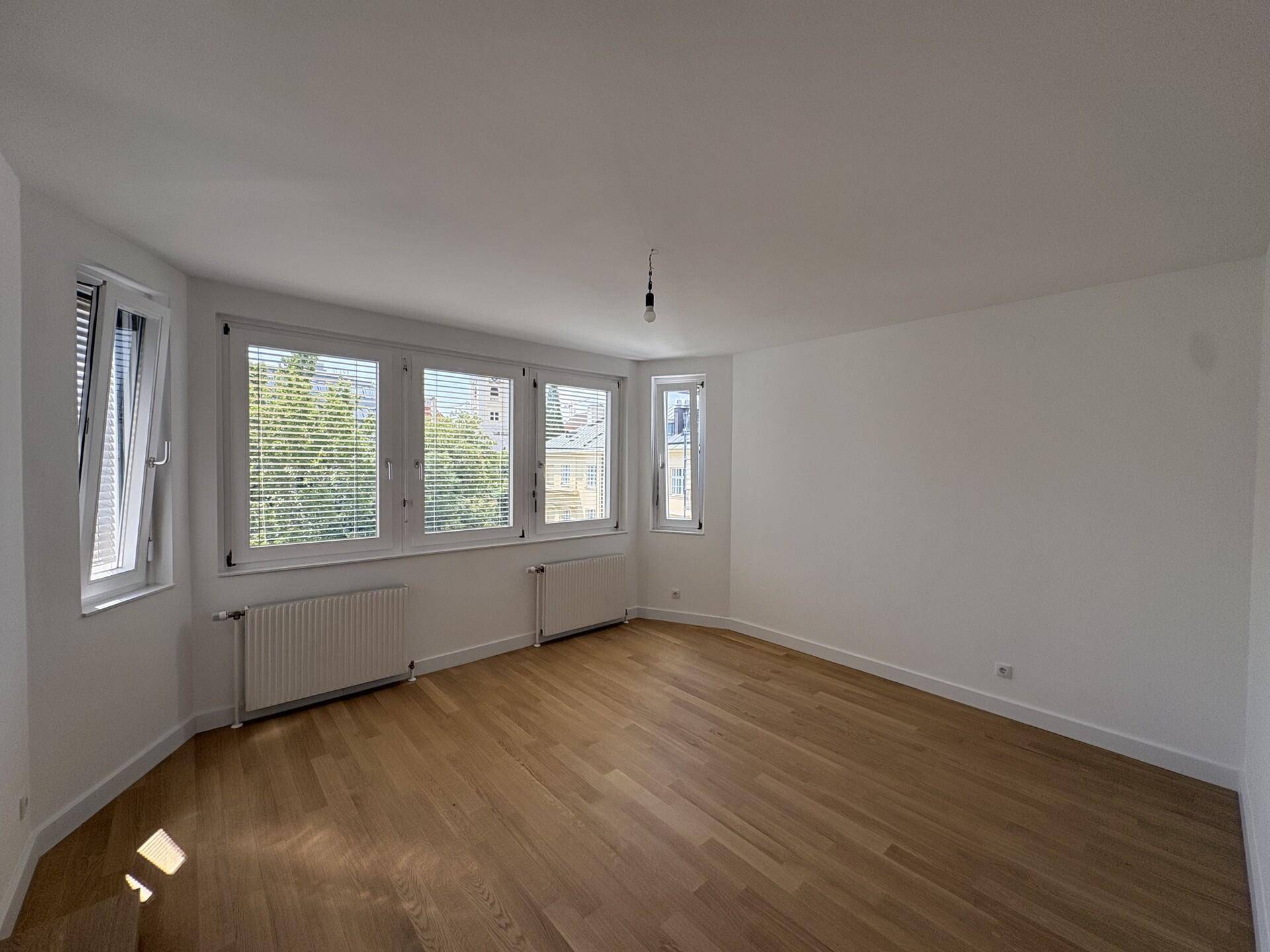 Helles Wohnzimmer mit Holzboden und mehreren Fenstern, die viel Tageslicht hereinlassen.