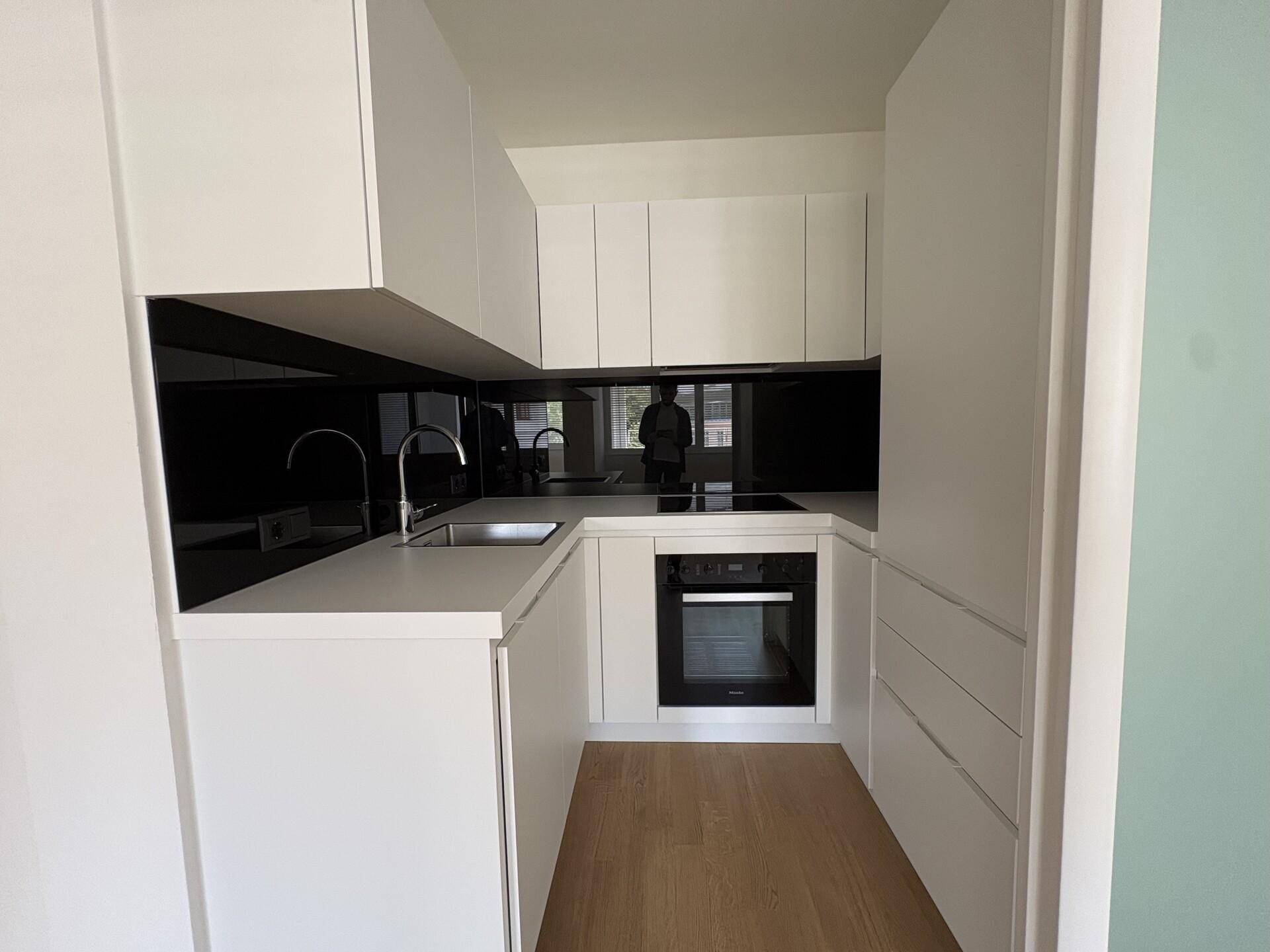 Kompakte, moderne Kochnische mit weißen Schränken, schwarzer Rückwand und integriertem Backofen.