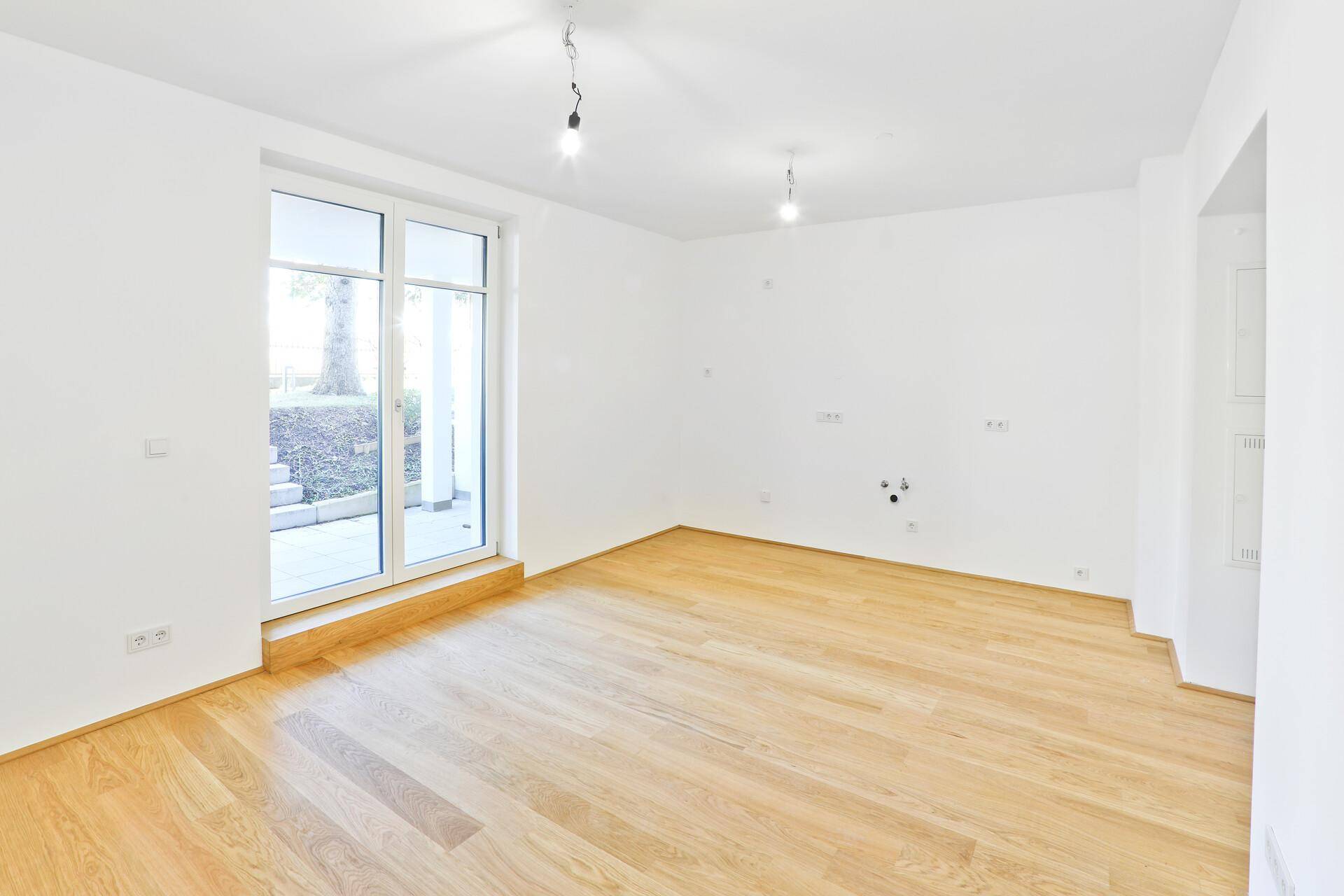 Heller, leerer Raum mit Holzboden und großen Fenstern zum Außenbereich.