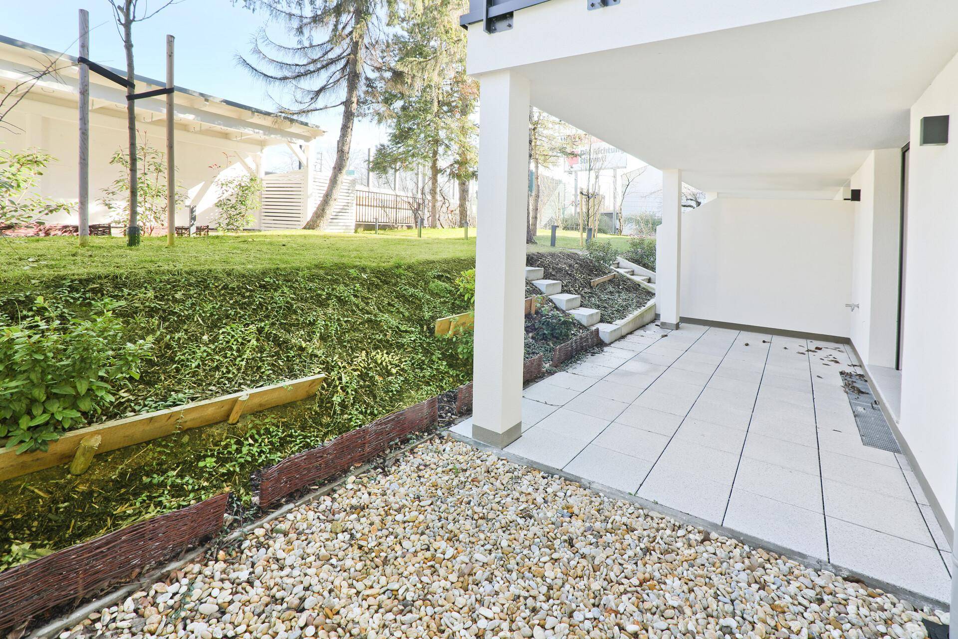 Überdachte Terrasse mit Fliesenboden und angrenzender Grünfläche mit Bäumen.