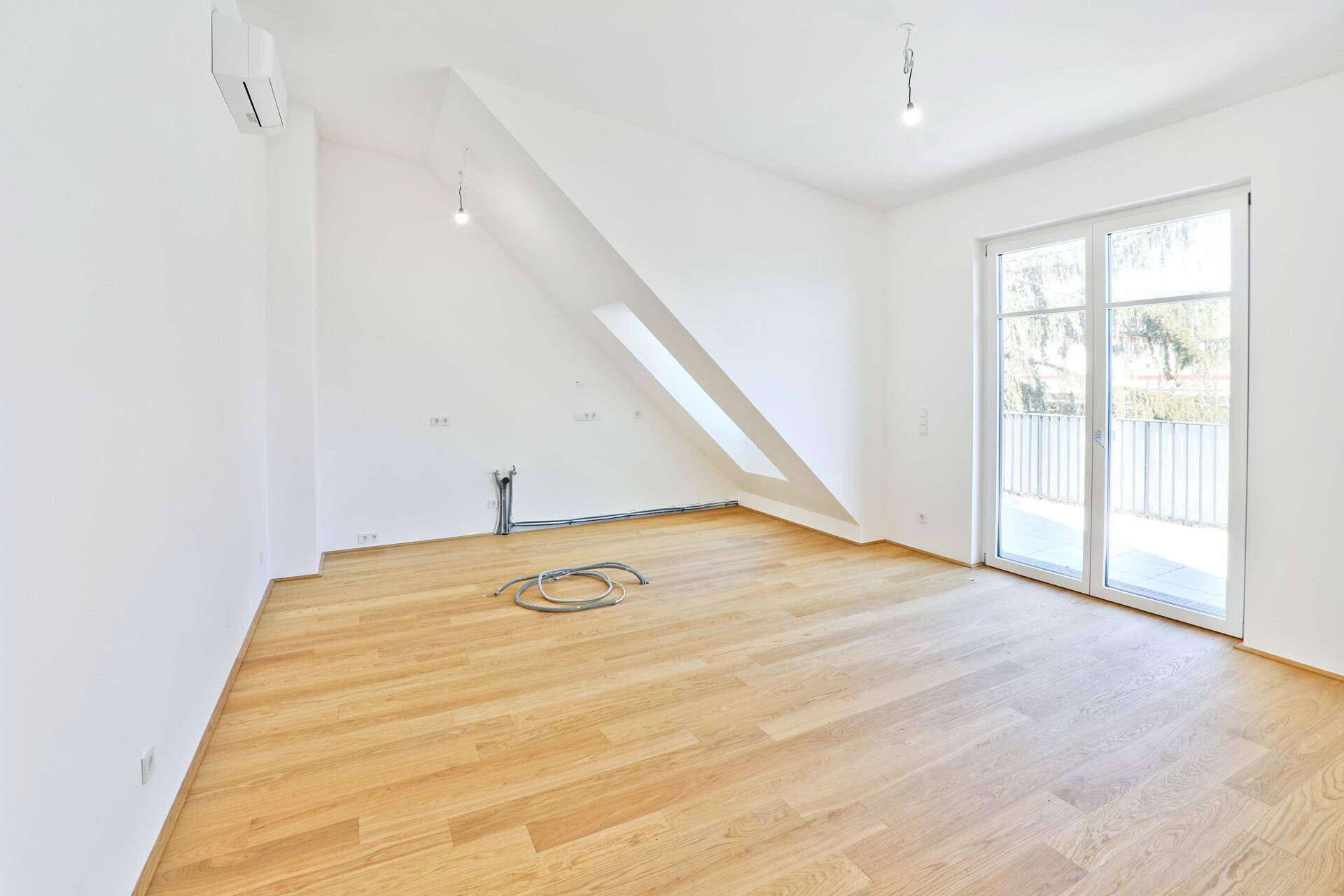 Geräumiger, heller Raum mit Holzparkett, Dachschräge und Zugang zu einem Balkon.