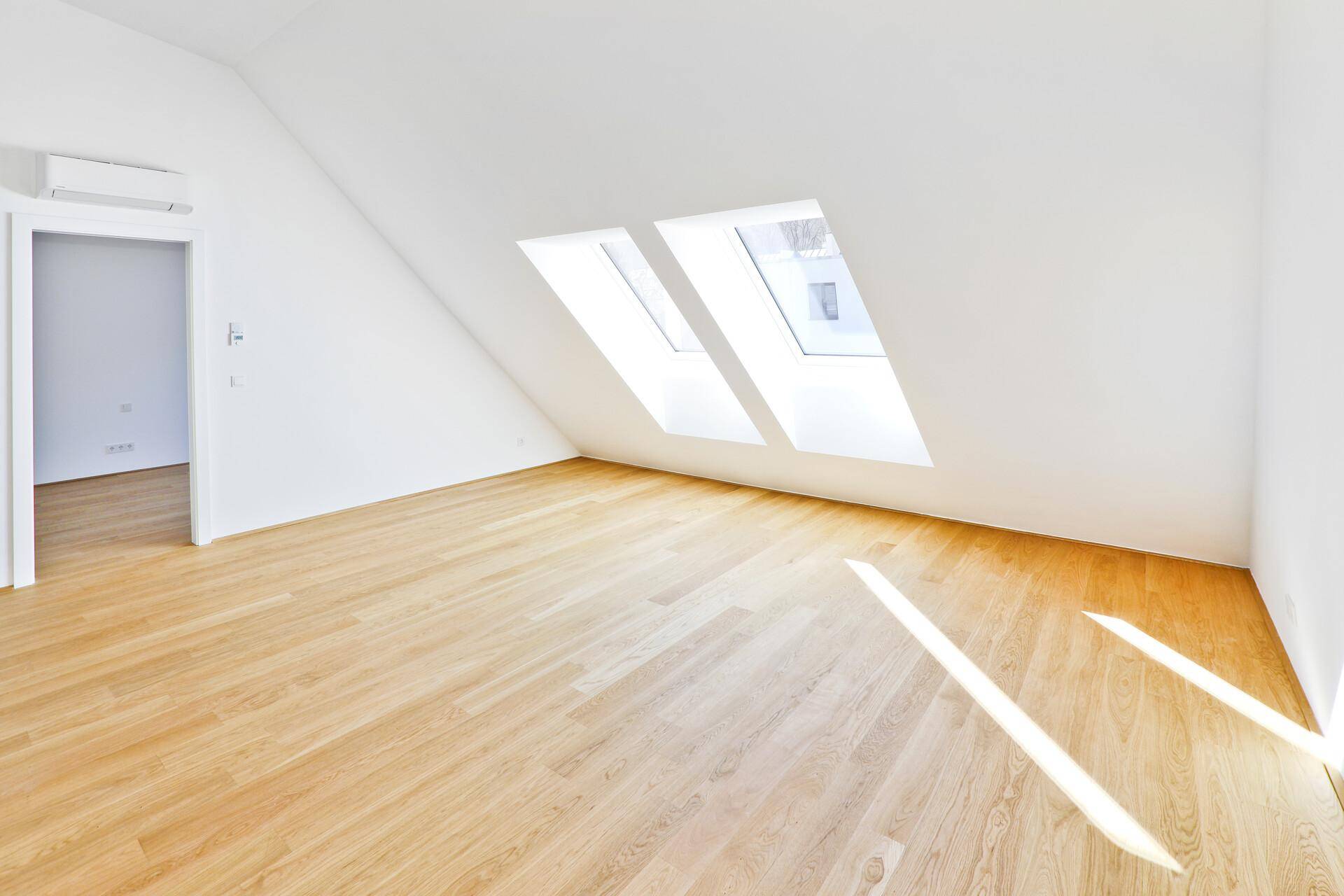 Heller Raum mit Holzparkett, zwei Dachfenstern und Sonnenlicht, das auf den Boden fällt.