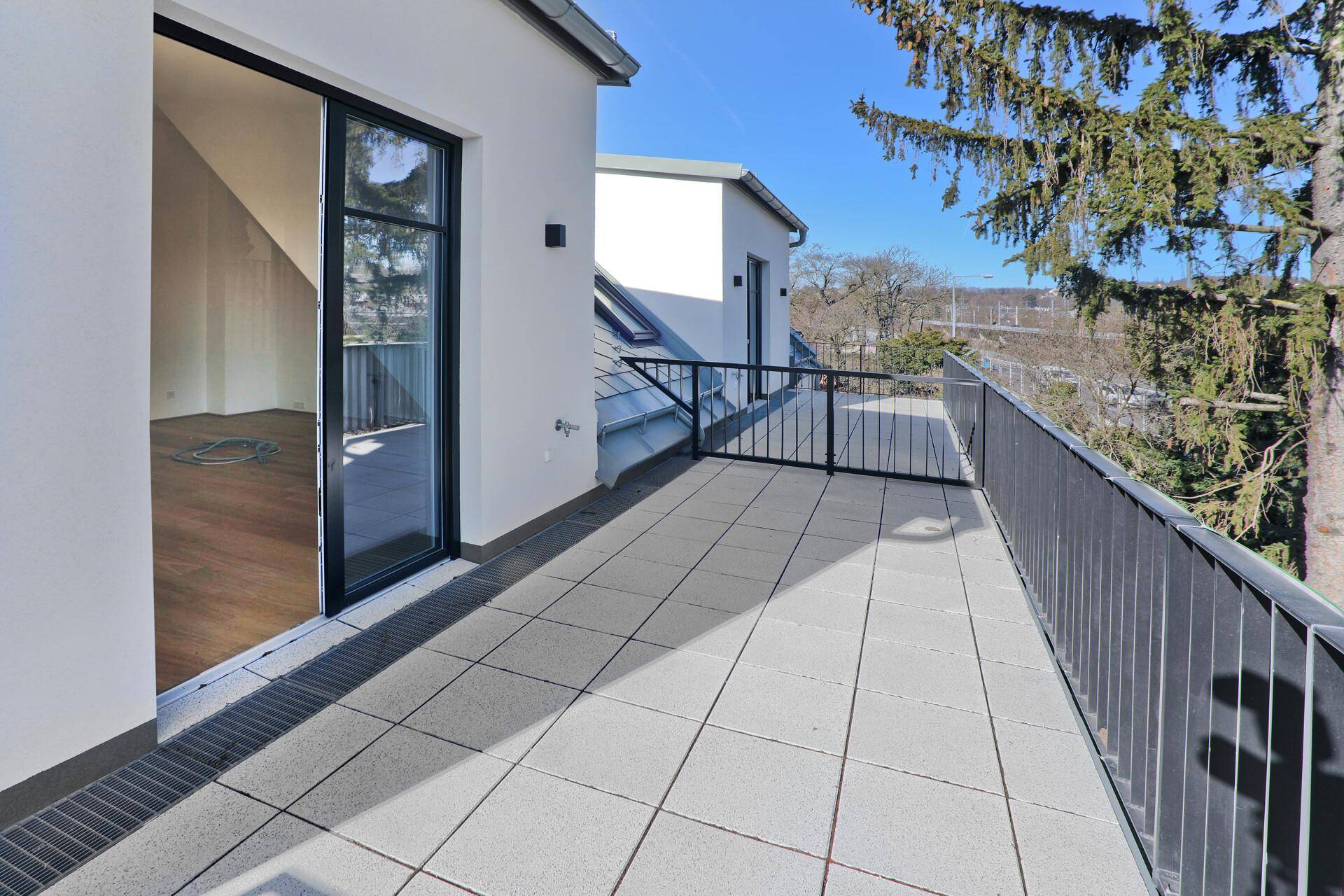 Langer Balkon mit Fliesenboden und Zugang zu einem hellen Innenraum, umgeben von Bäumen.