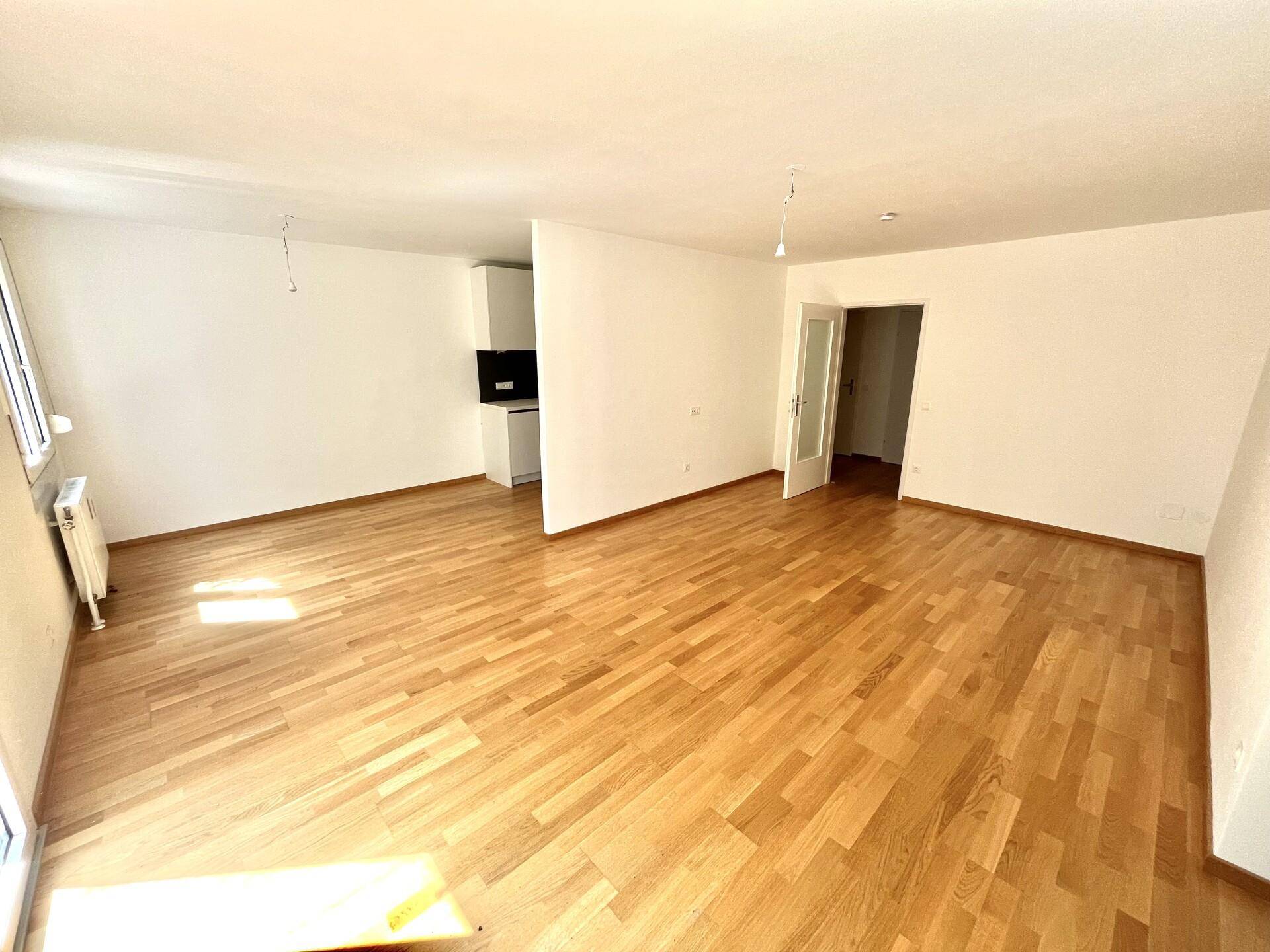 Heller, leerer Wohnbereich mit Parkettboden und offenem Übergang zur Küchenzeile.