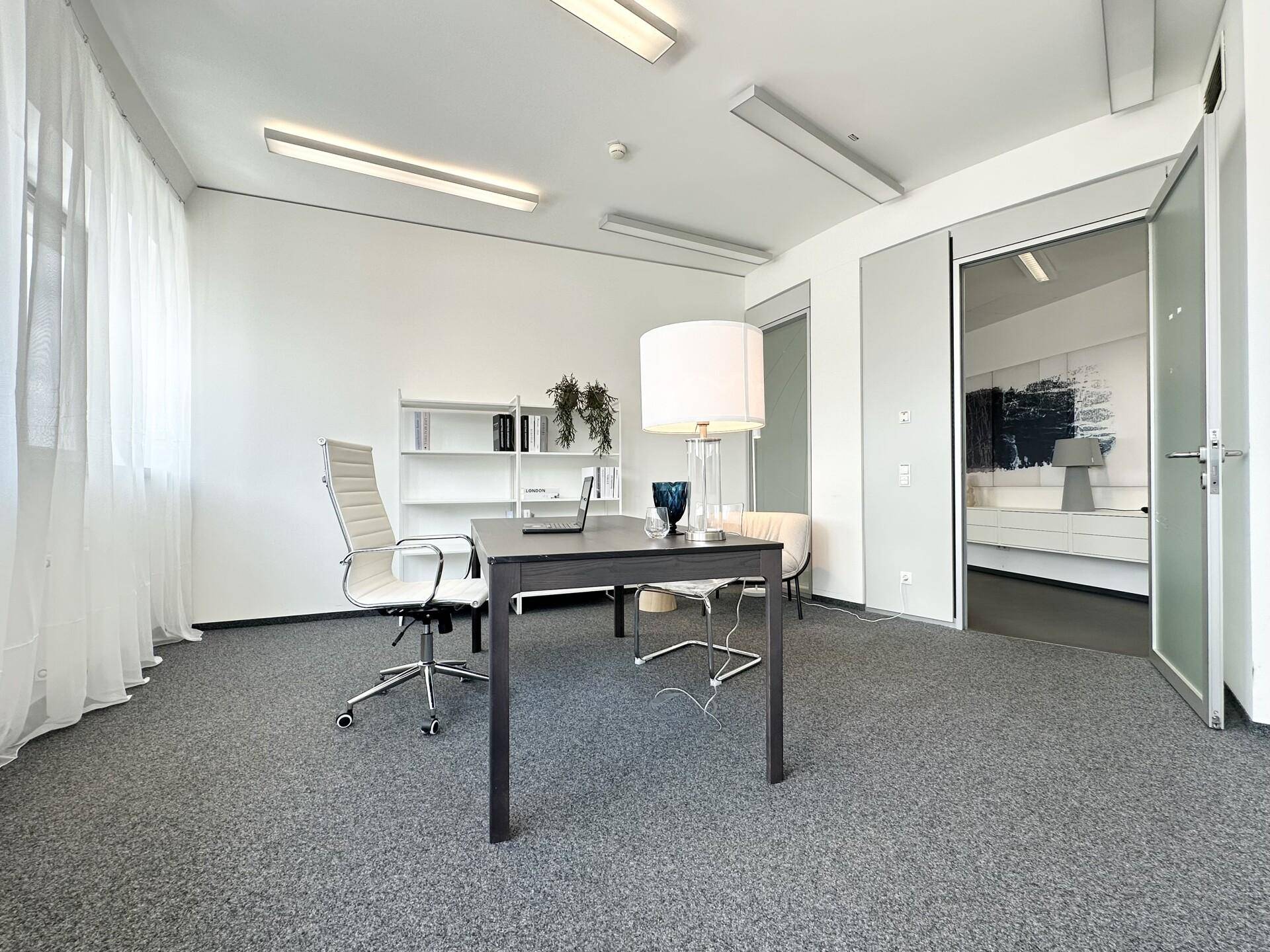 Helles Einzelbüro mit weißer Einrichtung, Schreibtisch und Blick in einen weiteren Raum durch eine Glastür.